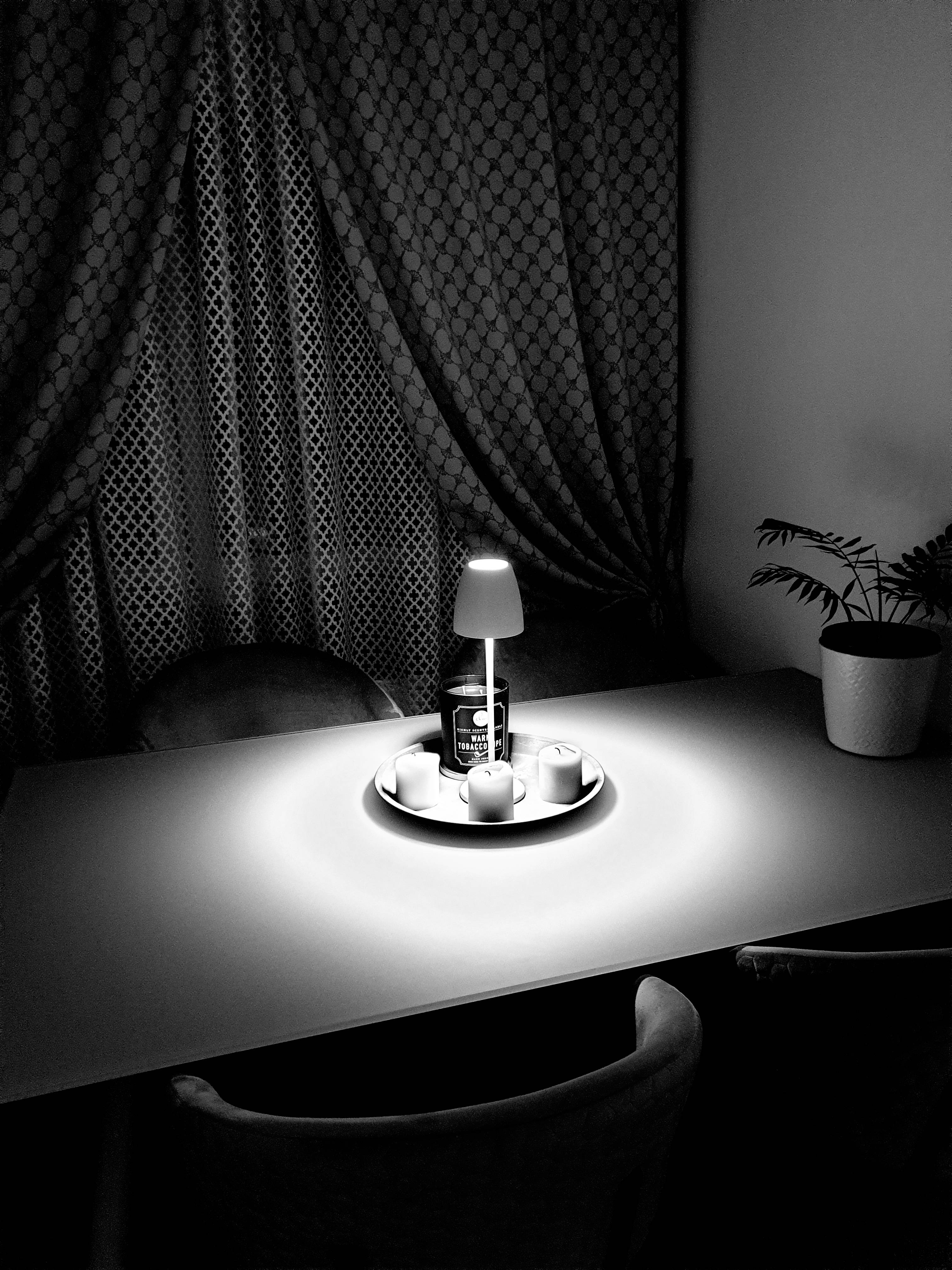 Ett bord med en liten lampa som lyser upp ett dryckeskärl och koppar. I bakgrunden en gardin och en växt.