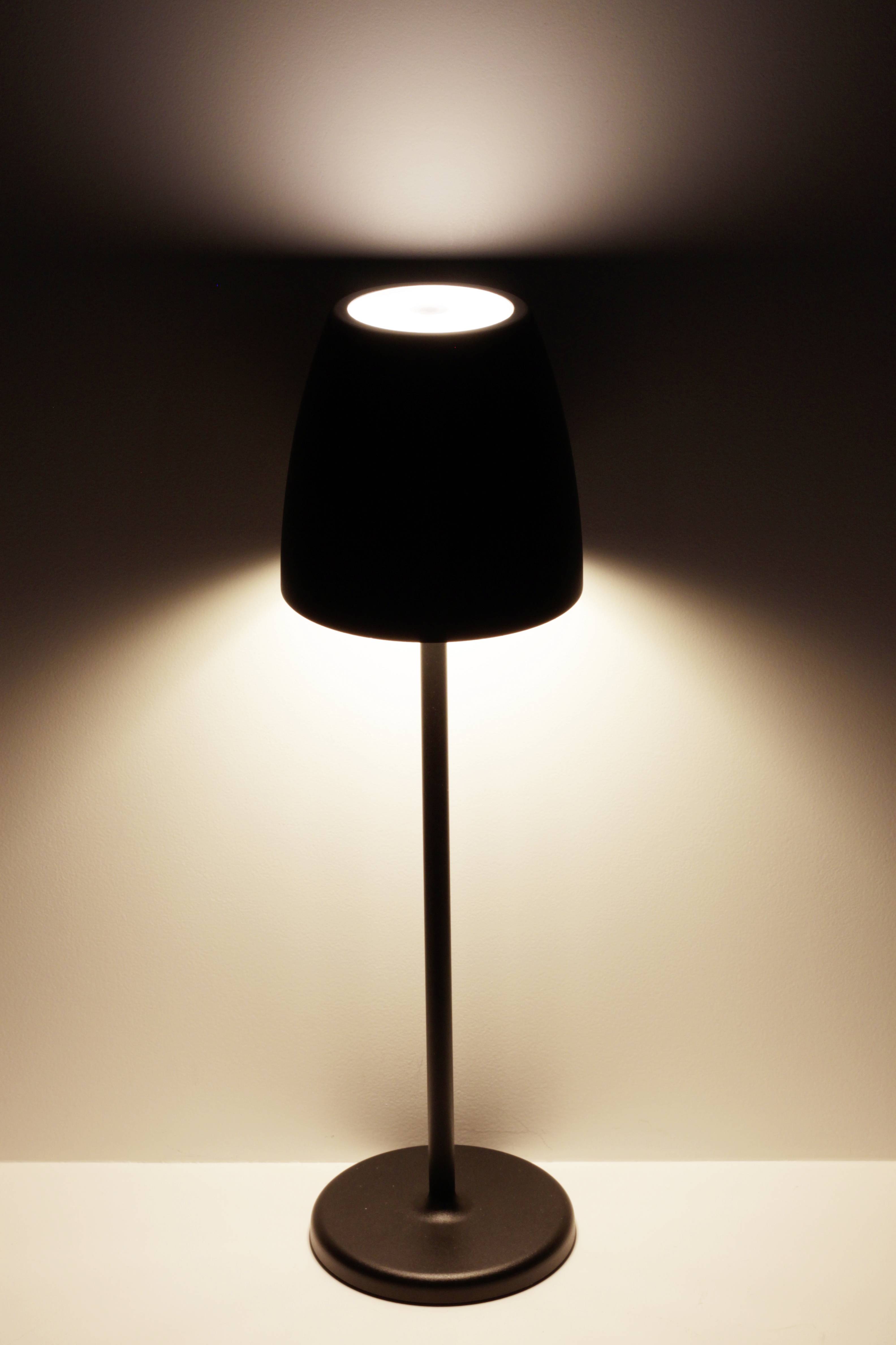 En bordslampa med en svart lampskärm står på en vit yta. Den lyser mjukt uppåt och nedåt.