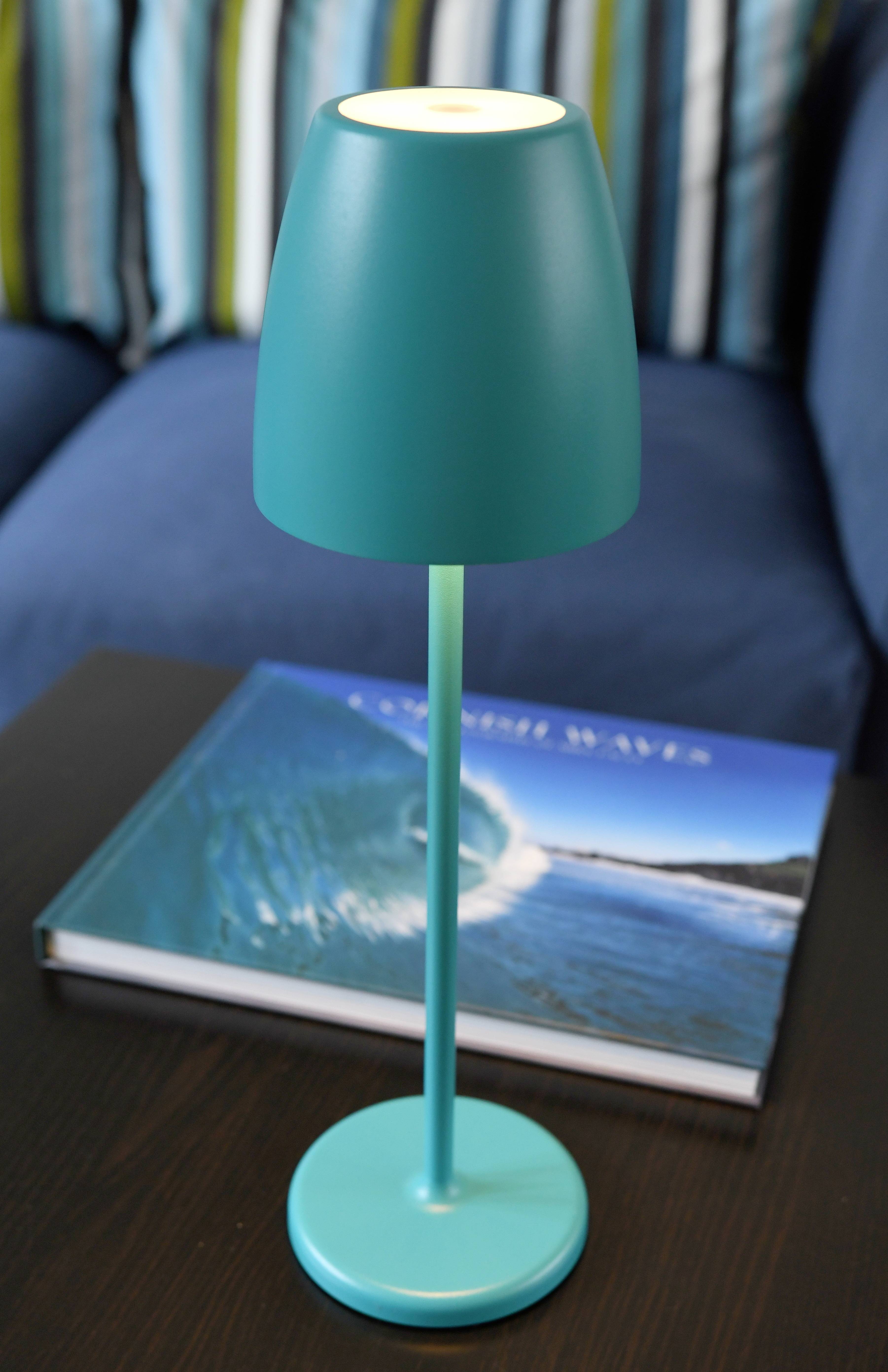 En turkos bordslampa står på ett mörkt träbord. Bredvid ligger en bok med titeln 'Crashing Waves' som visar en havsbild.