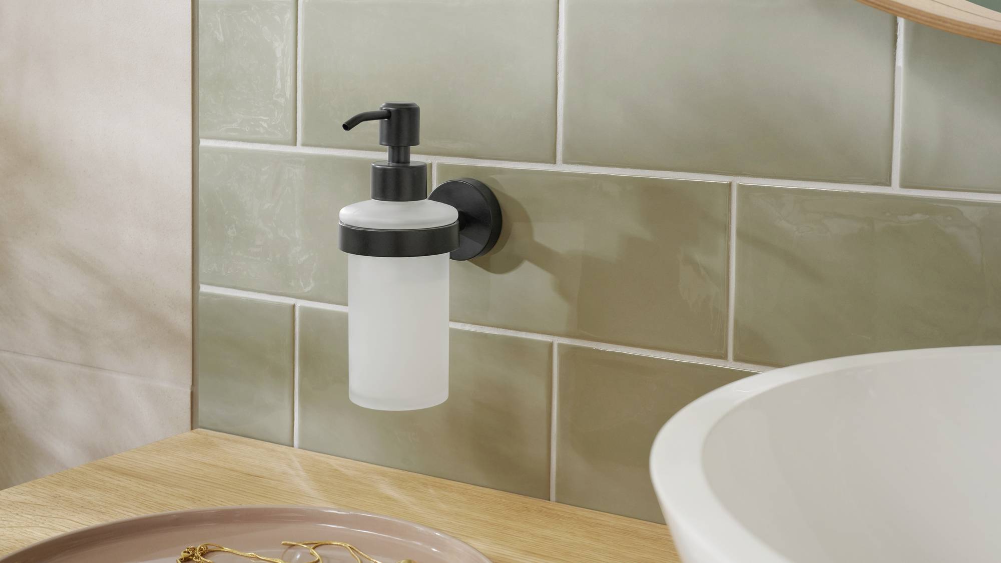 En tvålpump är monterad på en vägg med beige kakel bredvid en tvättställ. I förgrunden ligger ett bricka med smycken.