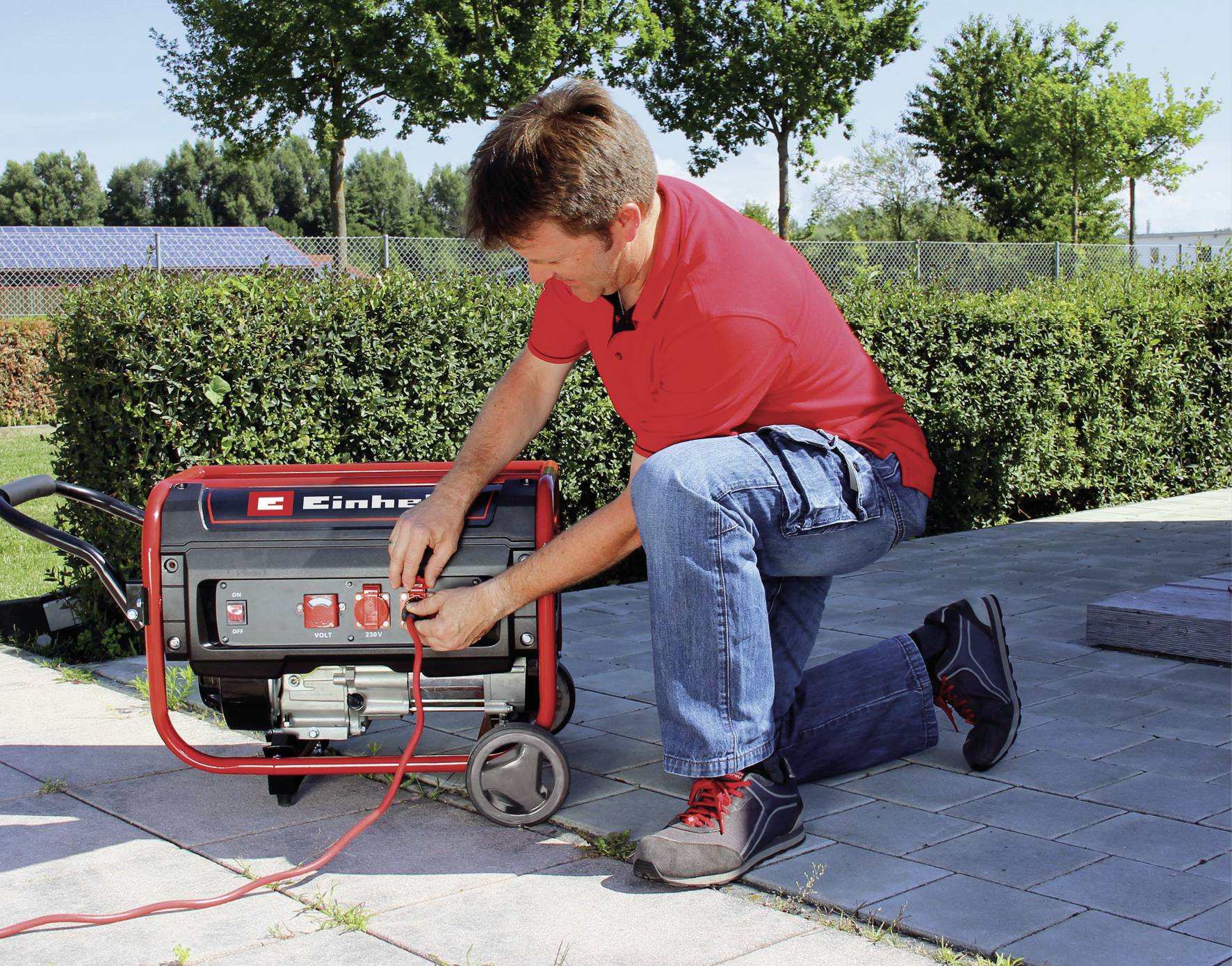 En man knäböjer och betjänar en röd-svart elgenerator utomhus, omgiven av växter och en stenlagd väg.