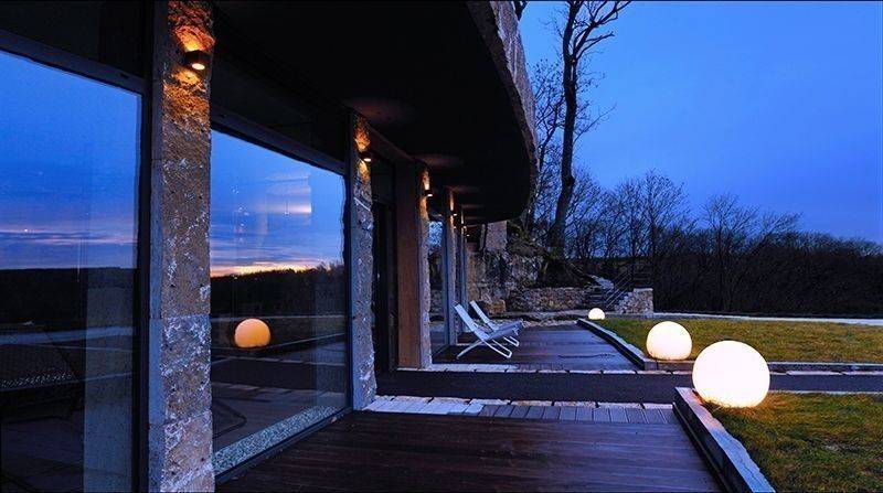Landskapsutsikt med en modern byggnad i förgrunden, stora fönster och upplysta klot längs en träterrass vid skymningen.