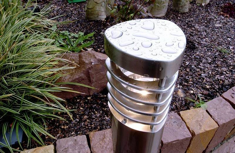 Trädgårdslampa av metall täckt med vattendroppar, omgiven av växter och stenar.
