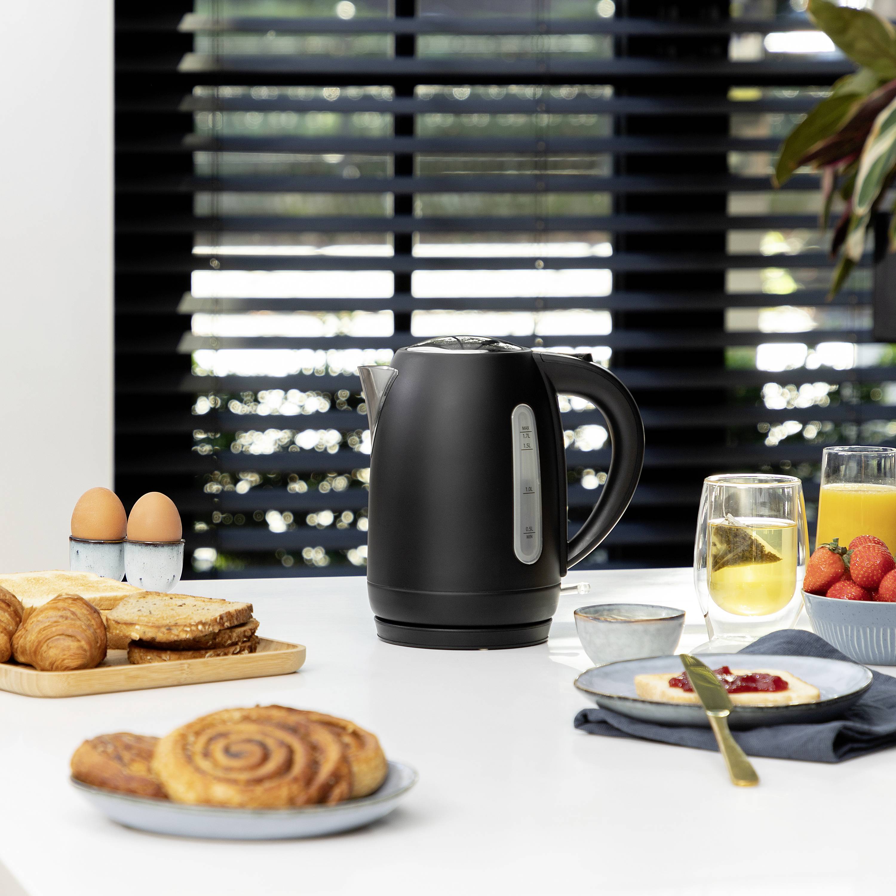En svart vattenkokare står på ett bord, omgiven av frukosttillbehör som croissanter, bröd, ägg och apelsinjuice.