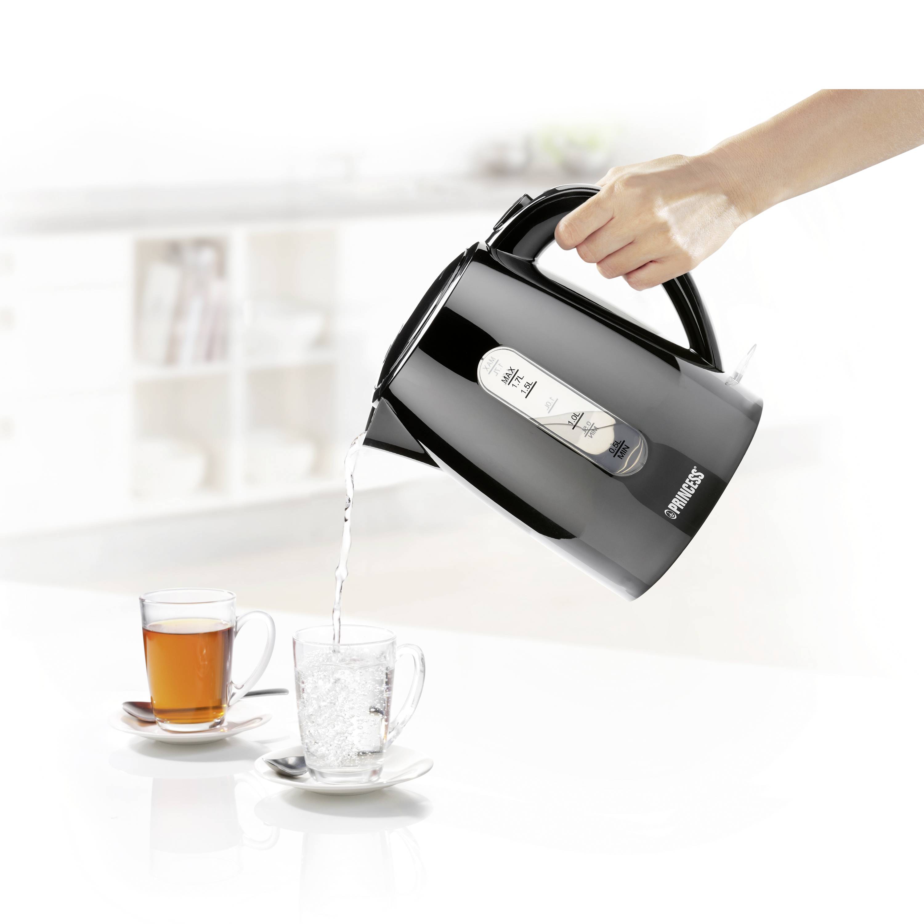 En hand håller en svart vattenkokare som häller vatten i ett glasmuggar. I bakgrunden står ytterligare en mugg med te.
