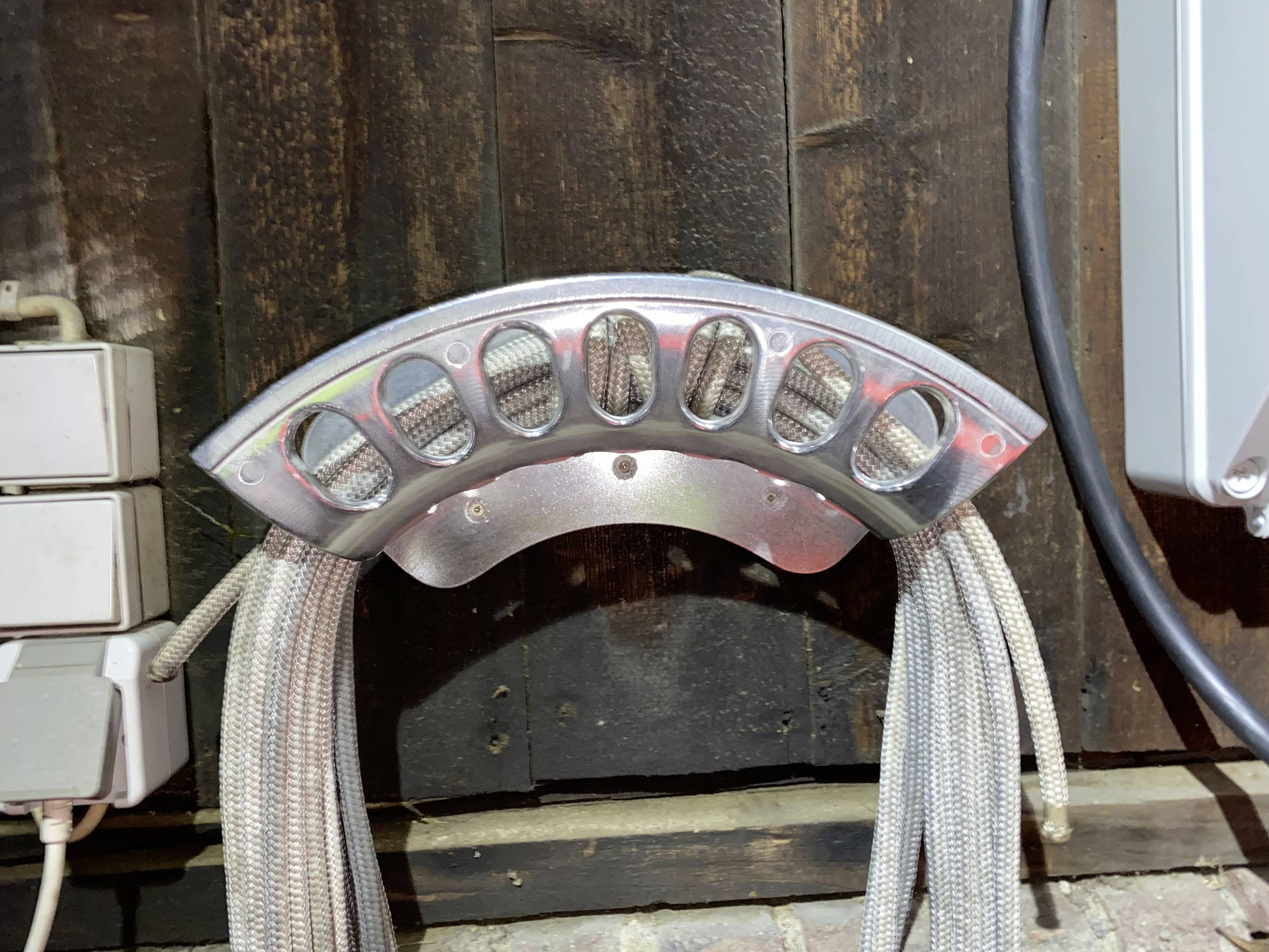 Metallhållare med flera grå kablar fästa vid en trävägg. Till vänster syns eluttag.