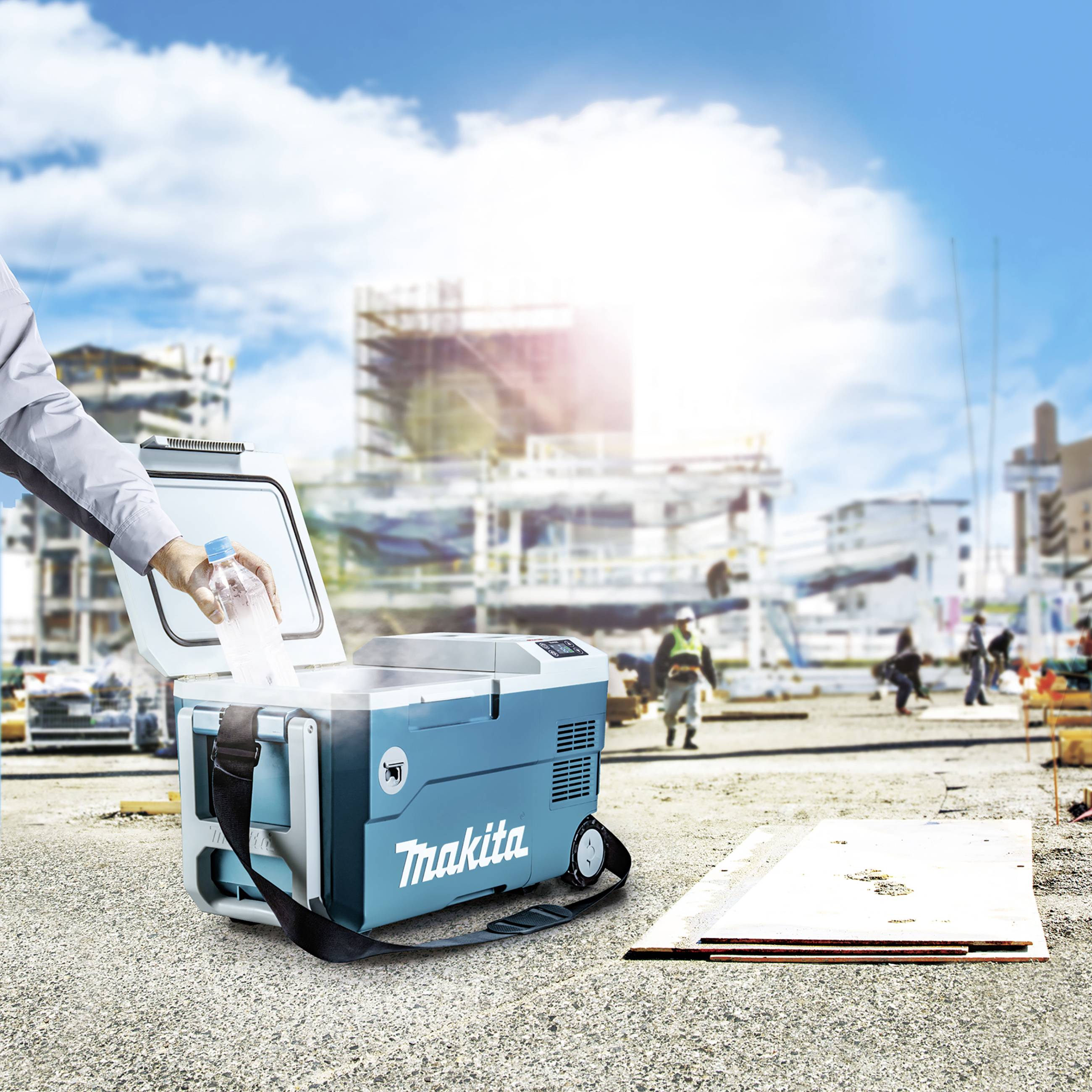 En person placerar en flaska i en blå kylväska med Makita-logotyp på en byggarbetsplats. Bakgrunden visar pågående byggnadsarbeten.
