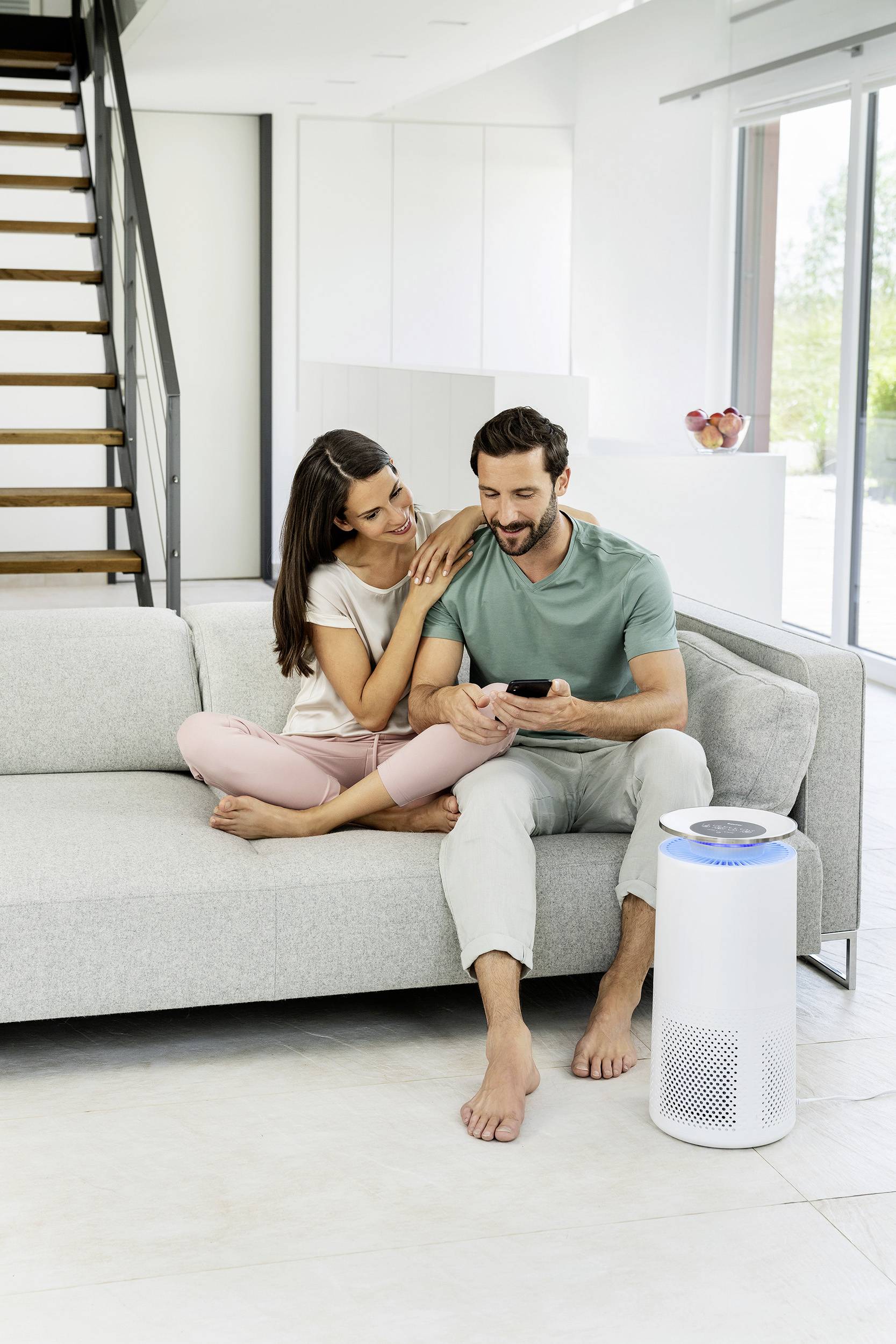 Ett par sitter på en soffa i ett ljust vardagsrum. De ler och tittar på en smartphone. Bredvid står en luftrenare.