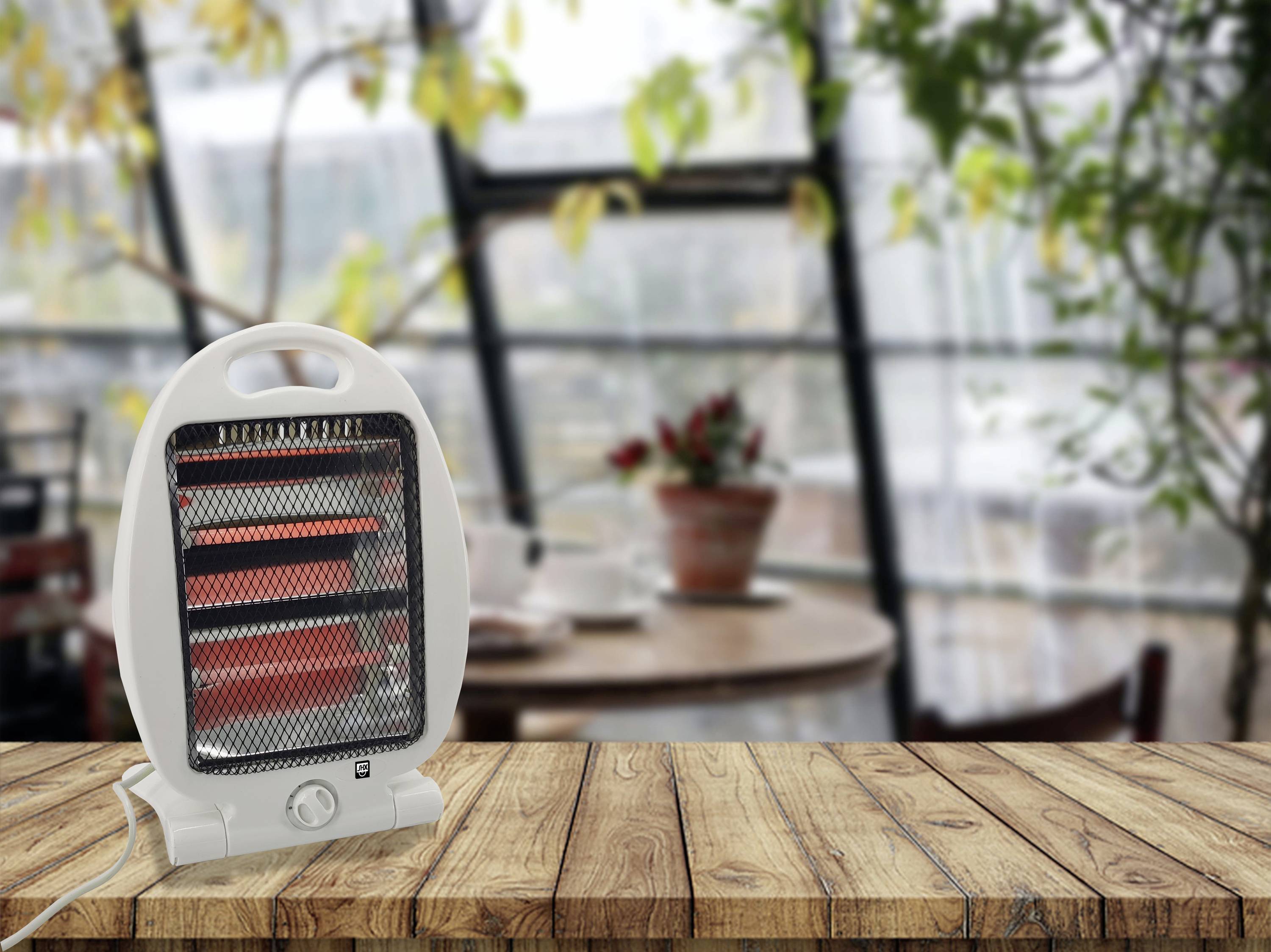 En liten elektrisk fläktvärmare står på ett träbord i ett mysigt rum med stora fönster och krukväxter.