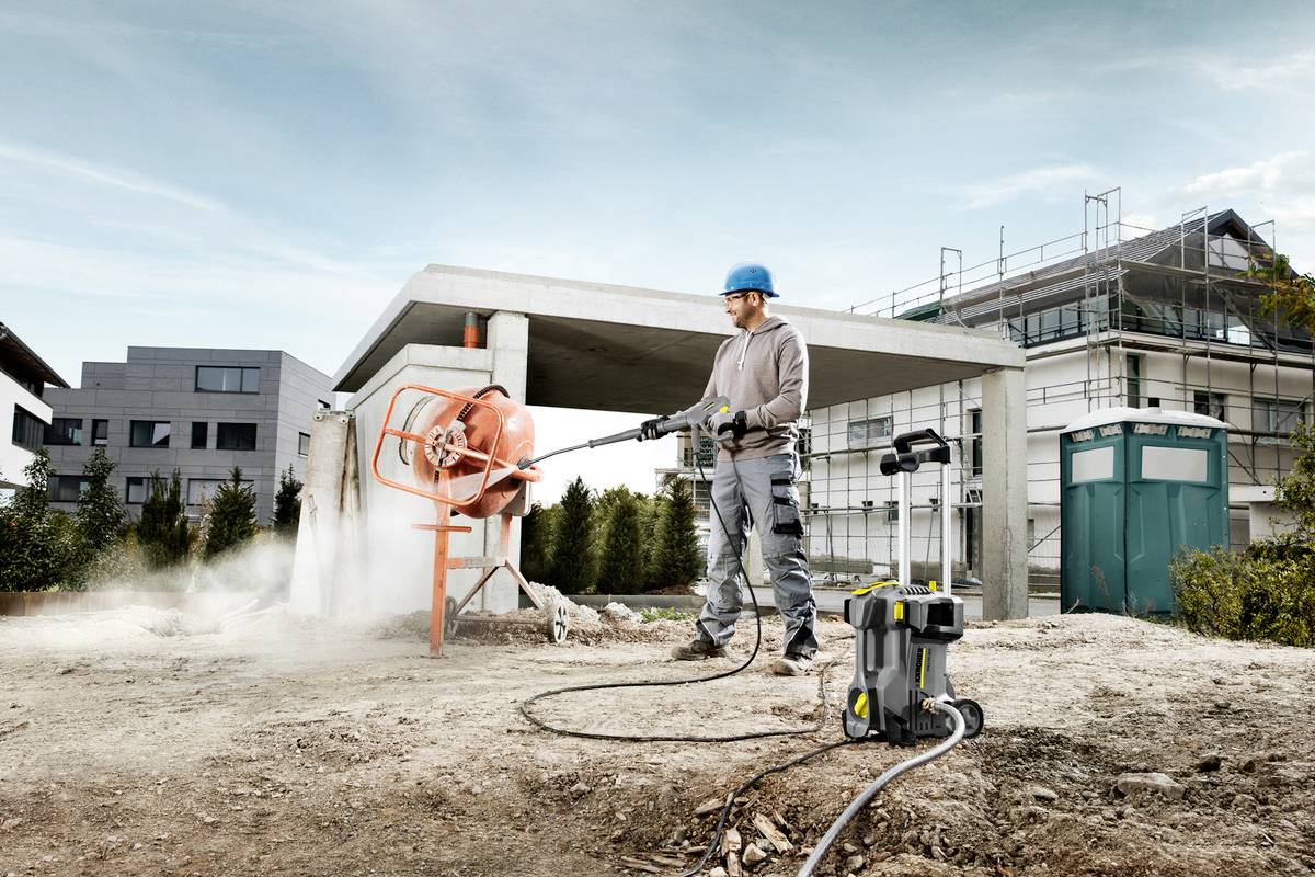 En byggnadsarbetare med hjälm sprejar vatten på en betongblandare på en byggarbetsplats. I bakgrunden syns byggnader och en ställning.
