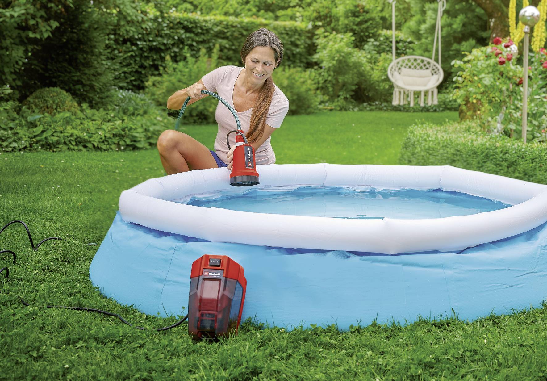 En kvinna tömmer en liten blå swimmingpool med en pump. Trädgården i bakgrunden är grön och frodig.