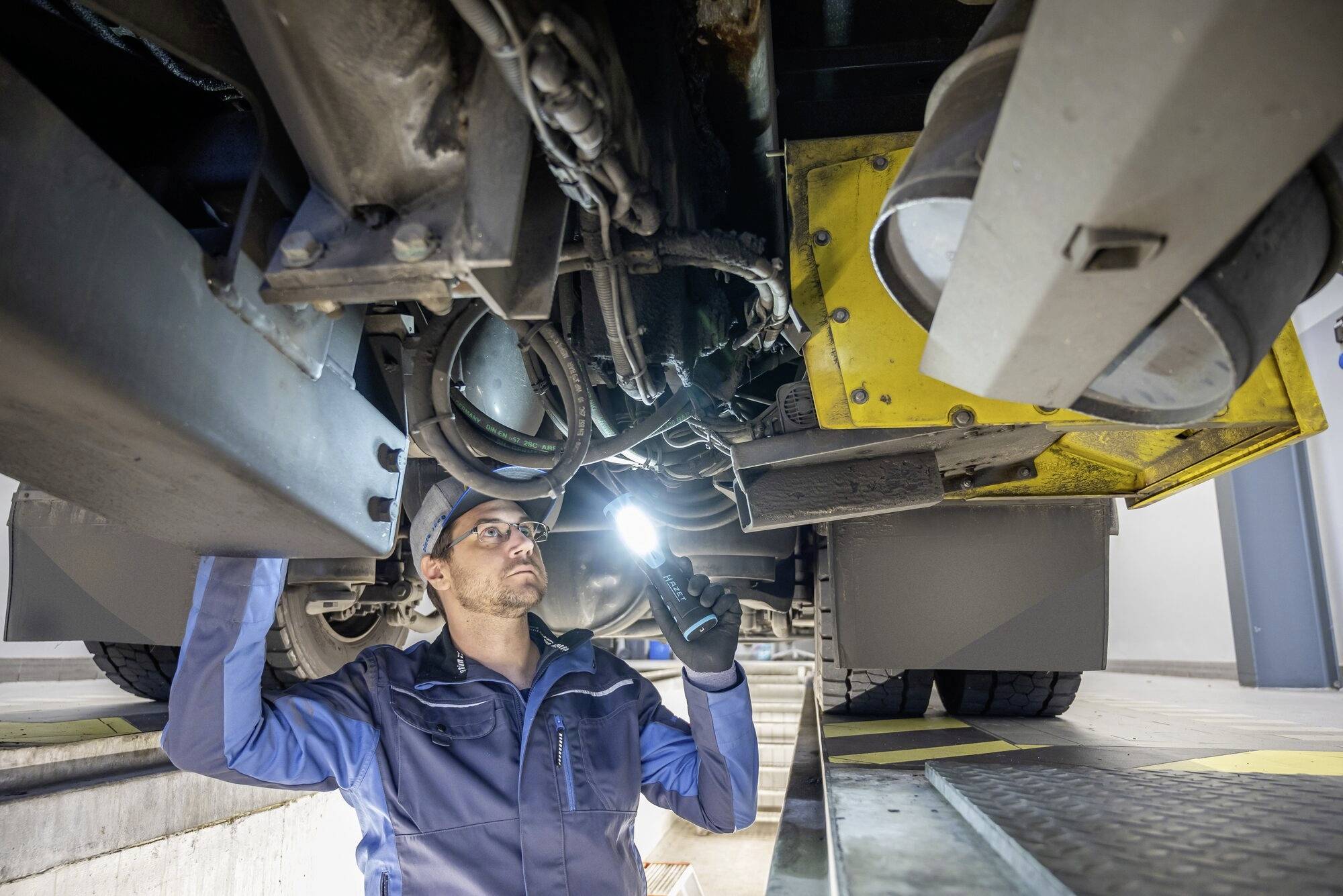 En mekaniker inspekterar underredet på ett fordon i en verkstad. Han bär arbetsklädsel och håller i en ficklampa.