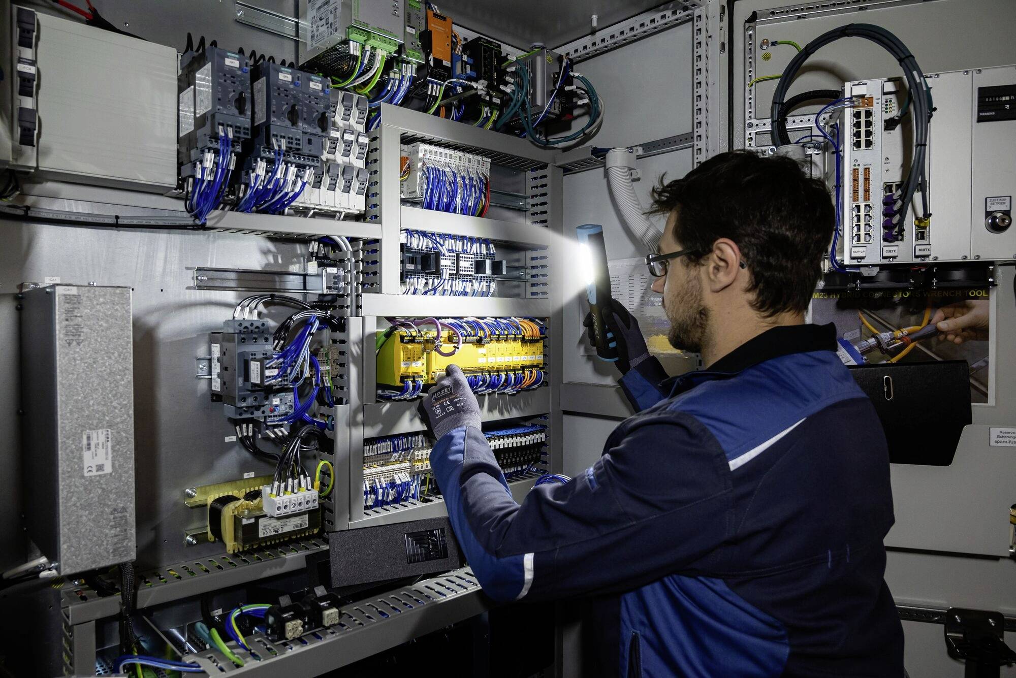 En tekniker kontrollerar elektroniska kretsar i en elskåp med hjälp av en ficklampa. Olika kablar och komponenter är synliga.