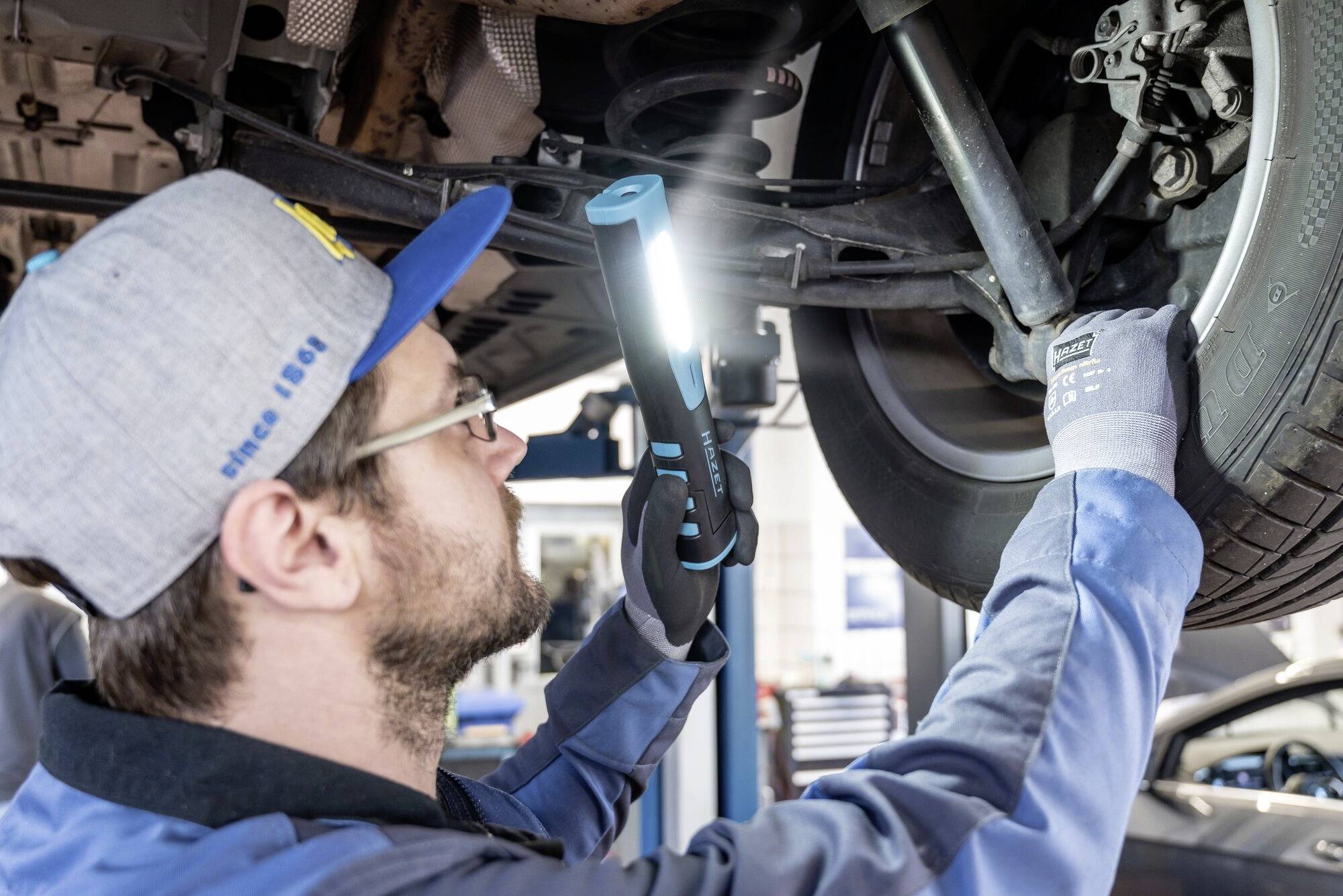 En person i arbetsklädsel inspekterar upphängningen på ett upplyft fordon med en lampa i en verkstad.