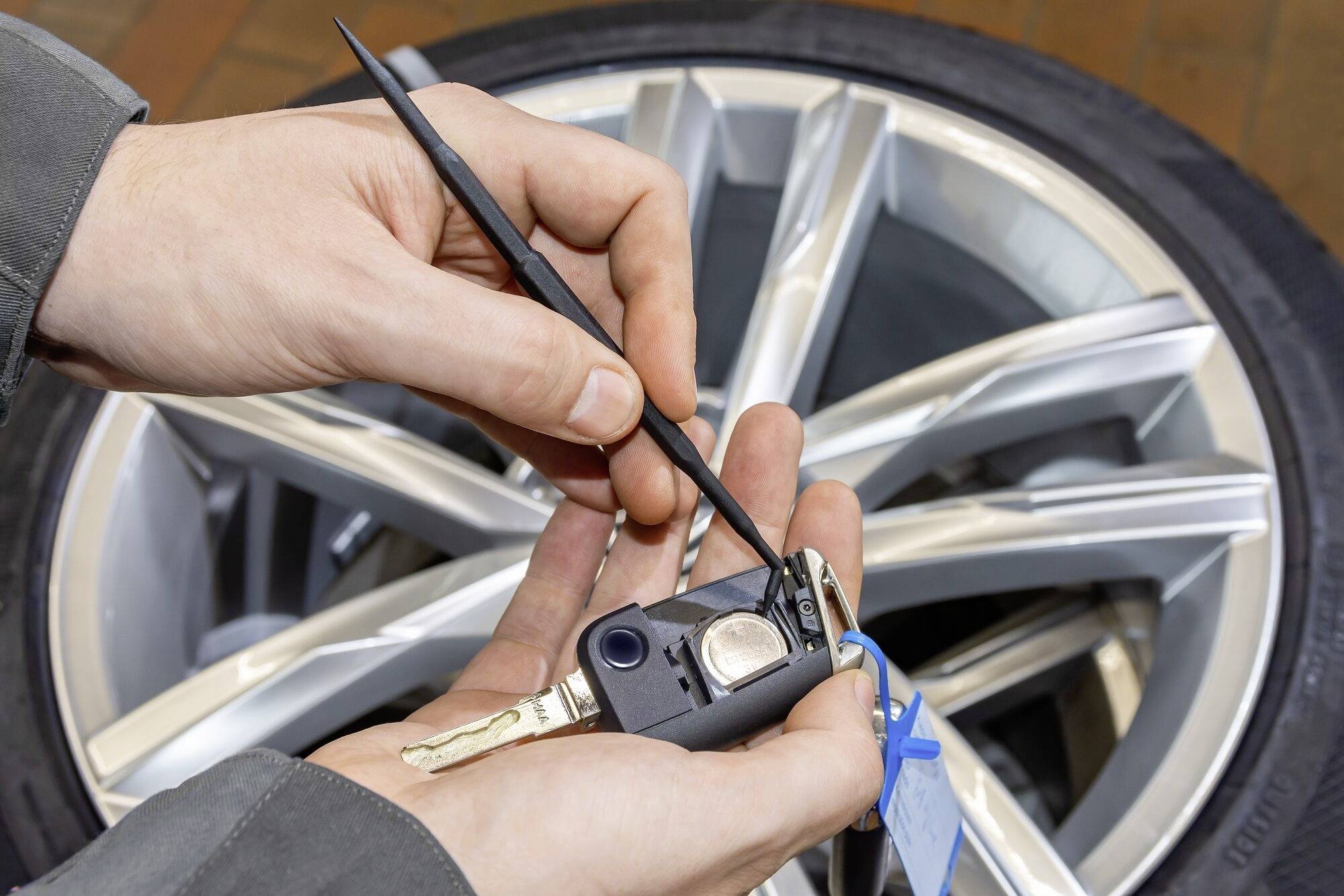 Händer håller en bilnyckel, ett däck i bakgrunden. En hand använder ett verktyg för att öppna nyckeln.
