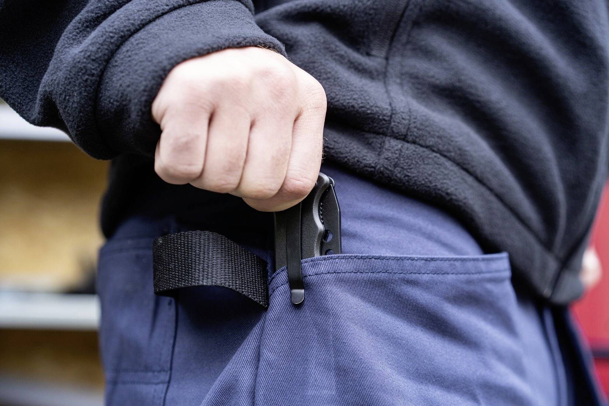 En person stoppar en svart fällkniv i sidofickan på sina blå byxor. Specifika detaljer på kniven är synliga.