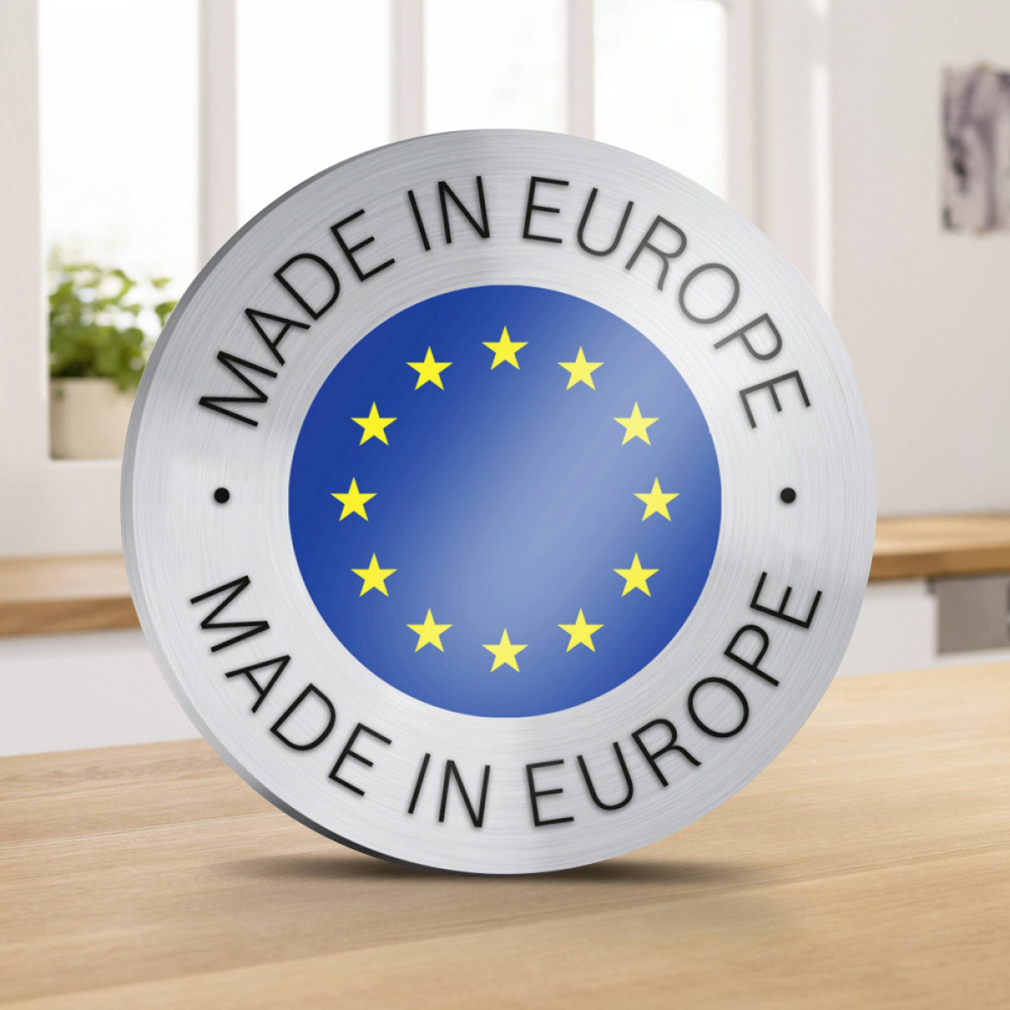 Rundlig emblem med EU:s stjärnring på blå bakgrund och texten 'MADE IN EUROPE'. Bakgrunden visar en suddig köksscen.