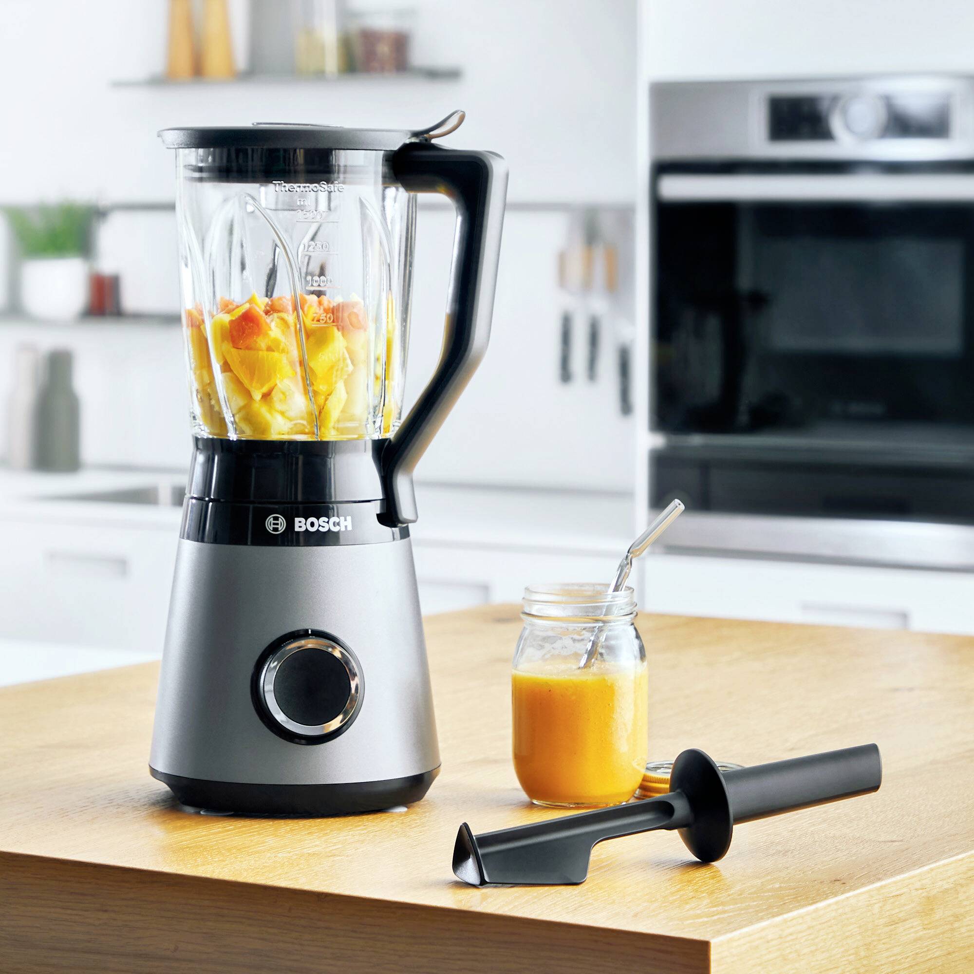 En mixer med skurna frukter på en träbänkskiva i ett modernt kök. Bredvid står ett glas med smoothie och en stöt.