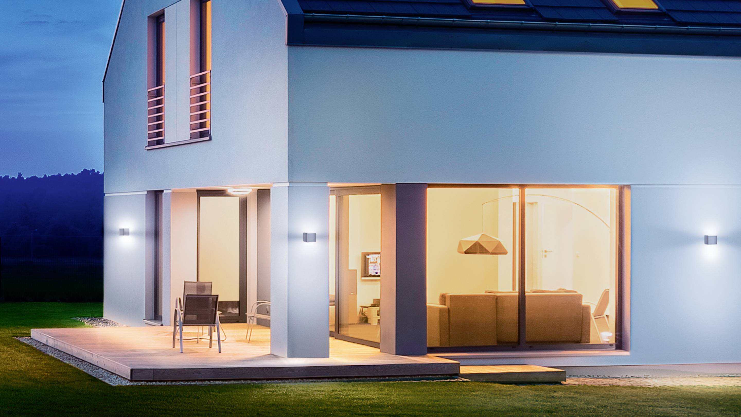 Modernt hus vid natten, upplyst inifrån, visar terrass med två stolar, fönsterfront med insyn i vardagsrummet.