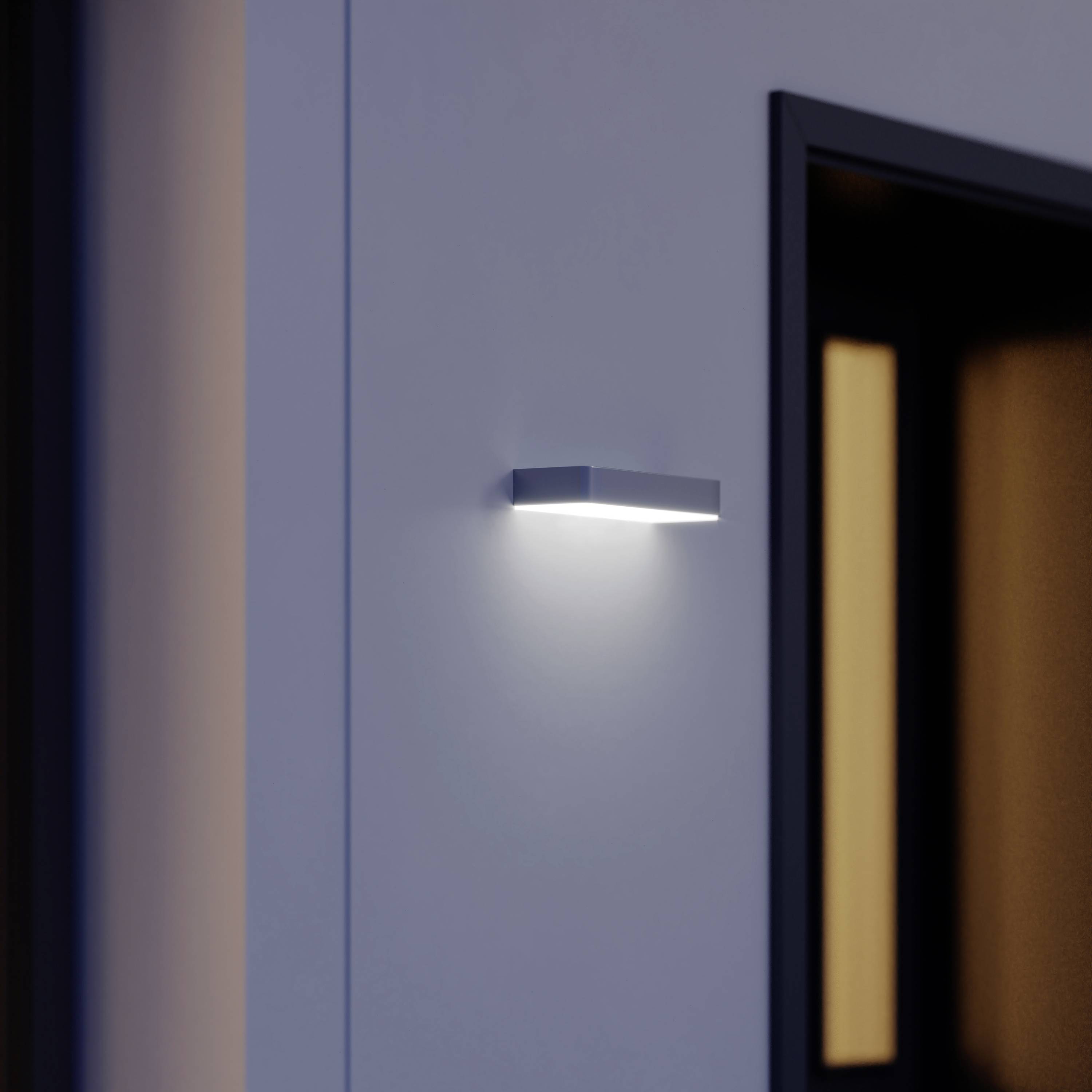 En modern vägglampa på en grå vägg kastar ett mjukt, diffust ljus som jämnt belyser området.