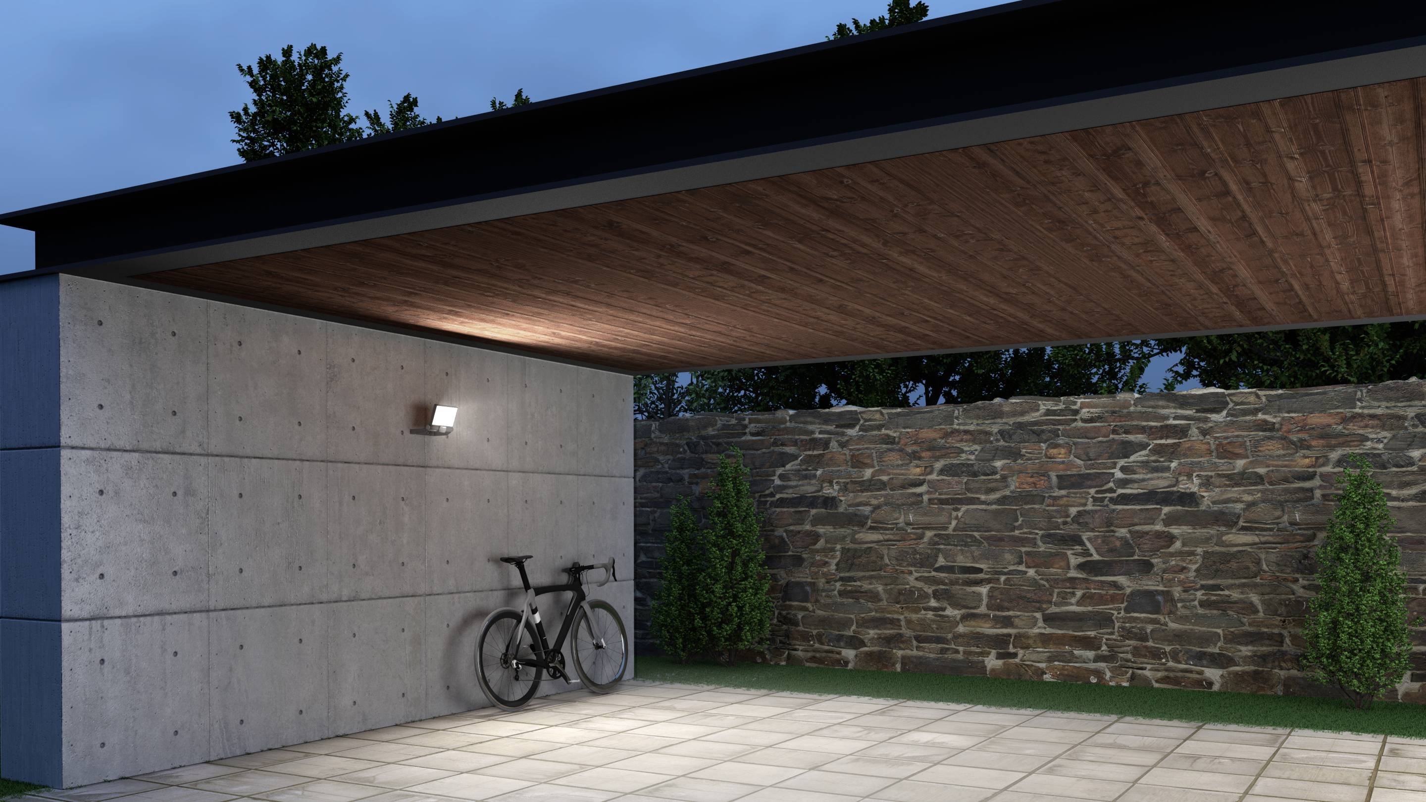Övertäckt carport med cykel mot betongvägg, bredvid stenmur och buskar i bakgrunden vid skymning.
