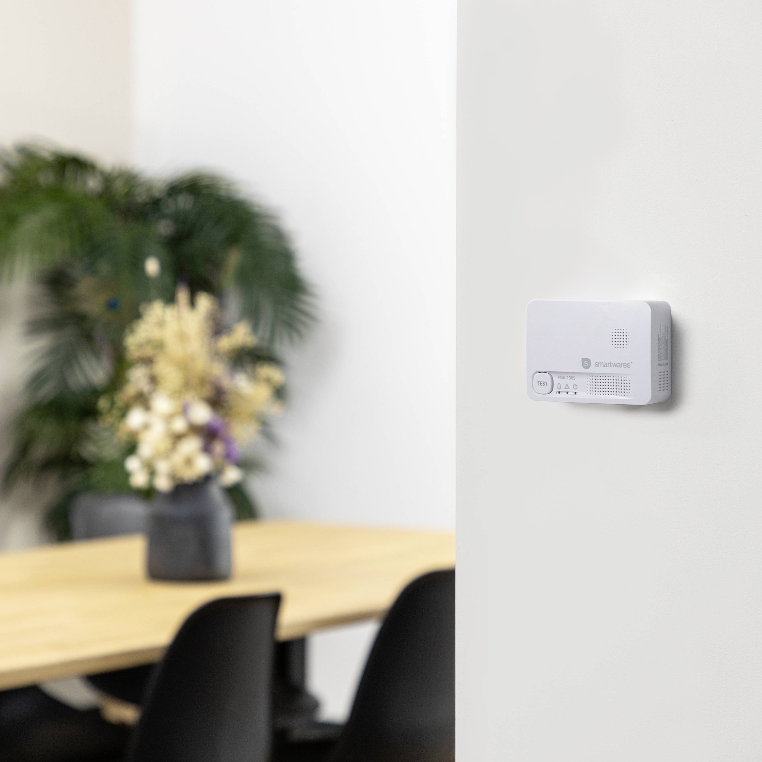 En modern termostat på en vit vägg. I bakgrunden, suddig, syns ett träbord med en blombukett och en växt.