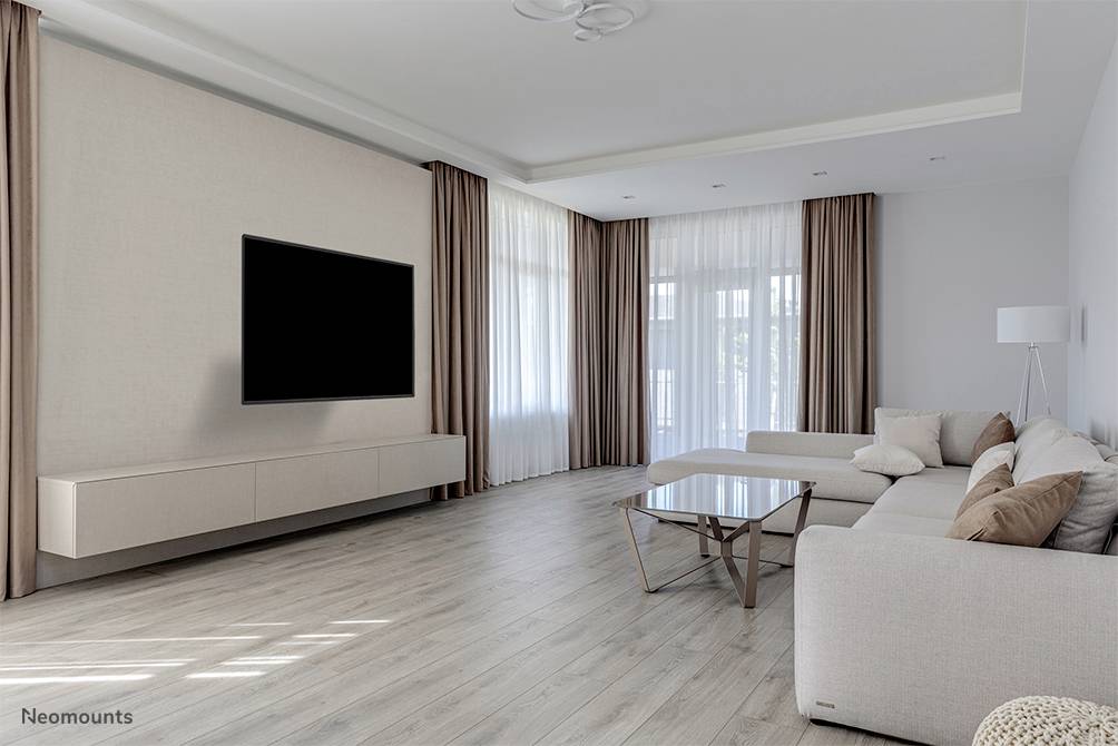 Helles vardagsrum med en modern soffa, glasbord och en stor flatskärms-TV på väggen. Stora fönster med långa gardiner.