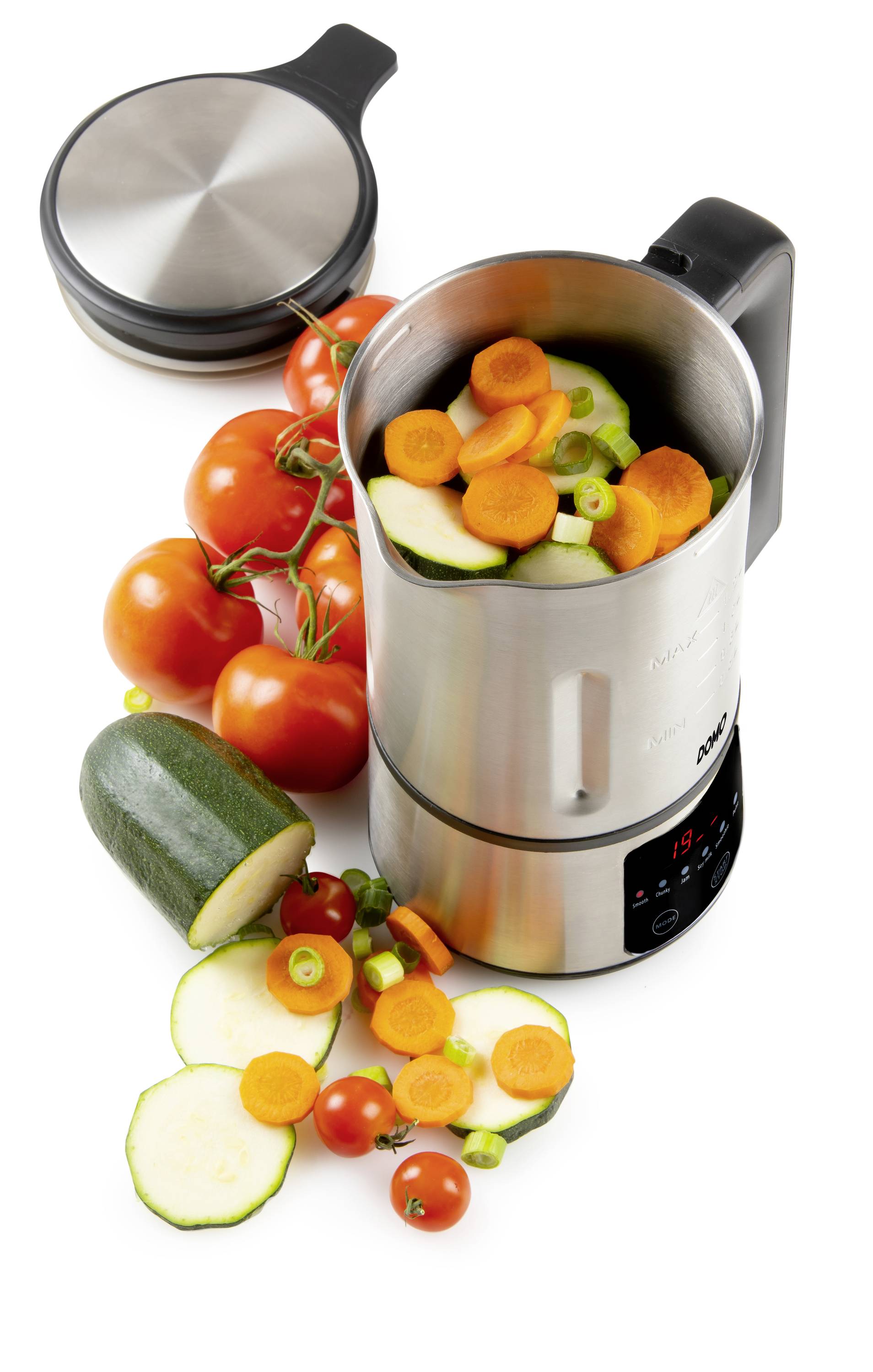 En rostfri mixer fylld med hackad grönsaker som morötter och zucchini. Bredvid ligger färska tomater.