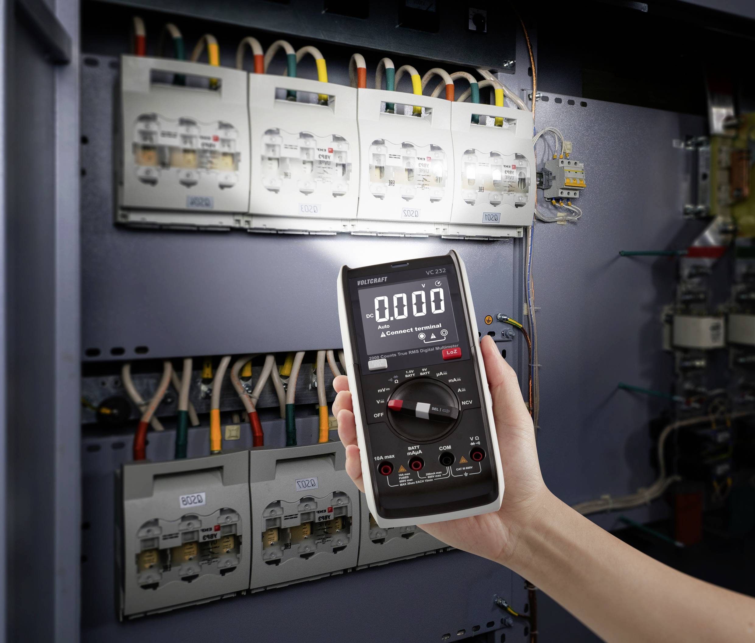 En person håller ett digitalt multimeter framför ett öppet elskåp med flera ledningsdragningar och säkringar.