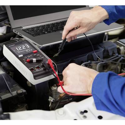 En person testar bilbatteriet med ett multimeter, medan en bärbar dator ligger öppen bredvid.