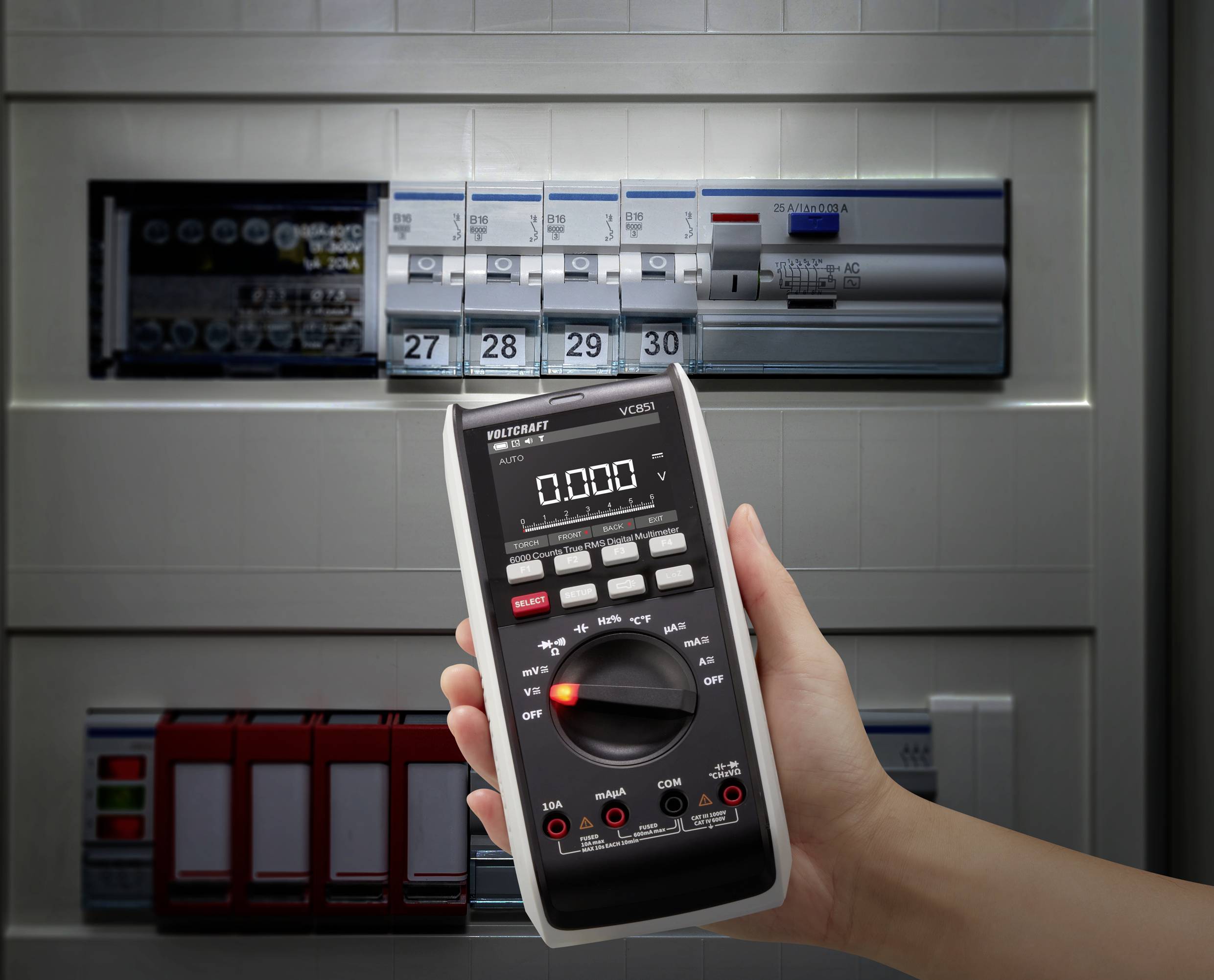 En hand håller ett multimeter som visar 0.00, framför en öppen elcentral med strömbrytare och säkringar, vilket symboliserar en elektrisk kontroll.