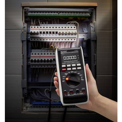 En hand håller ett digitalt multimeter framför en öppnad elcentral för att mäta elektriska värden.
