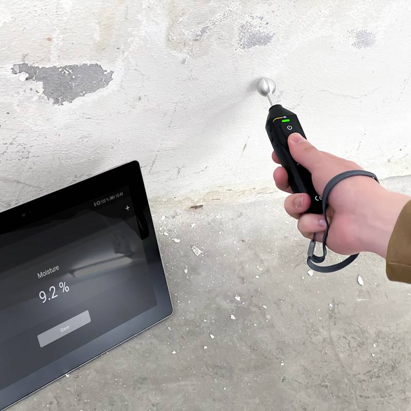 En hand håller ett fuktmätningsinstrument mot en vägg. En bredvidliggande surfplatta visar '9,2 %'.