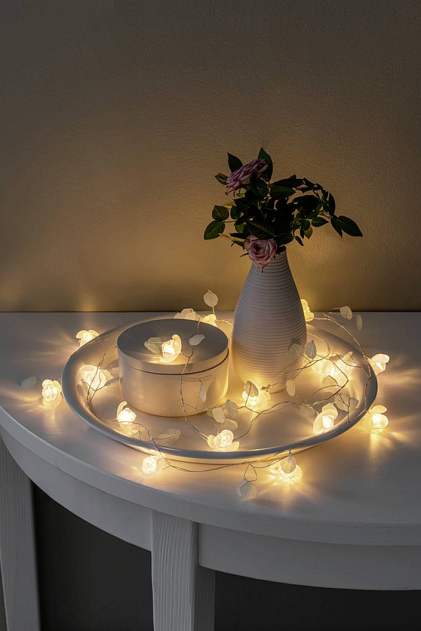 Ljusslinga i blomform på ett bord, arrangerad runt en vas med rosor och en rund ask. Varmt, stämningsfullt ljus.