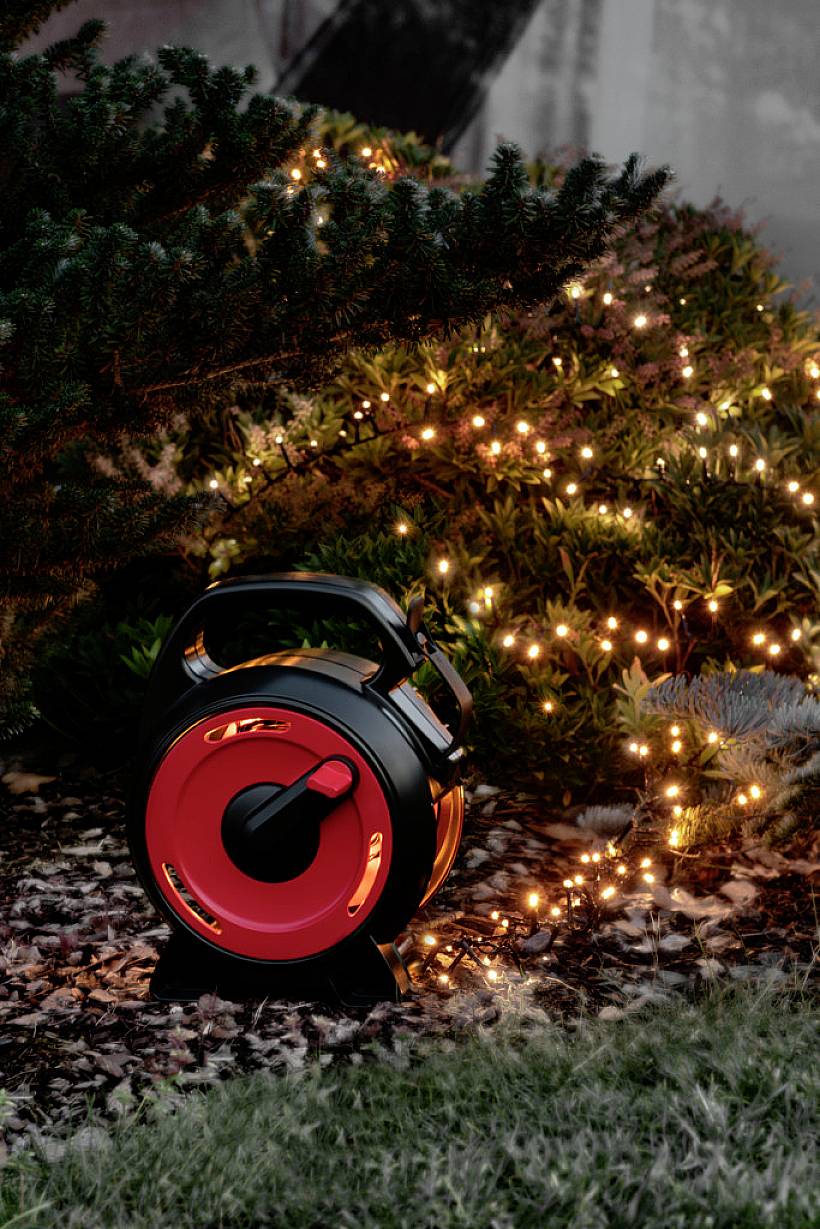En kabeltrumma med röd kant ligger i gräset och förser blinkande ljusslingor som lyser upp en buske under natten.