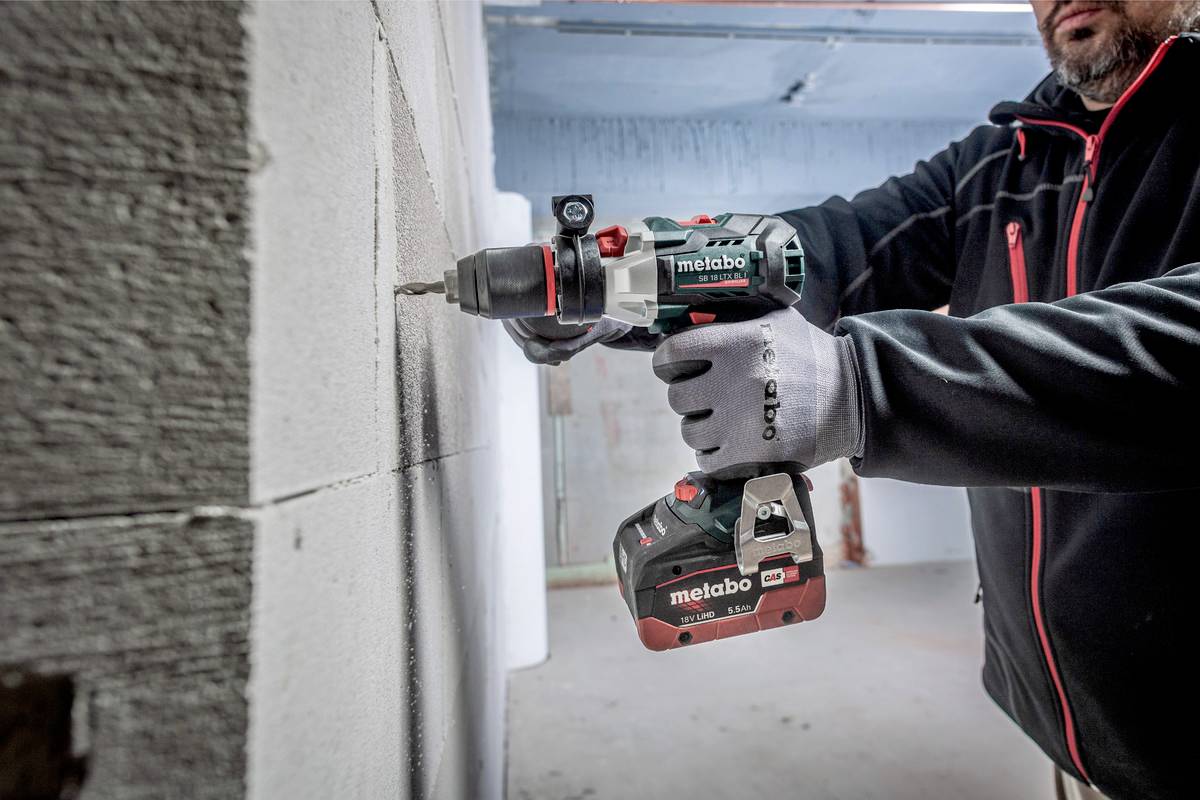 En person borrar i en vägg med en Metabo sladdlös borr. Personen bär arbetsklädsel och handskar, och rummet verkar vara i ett obearbetat byggnadsstadie.