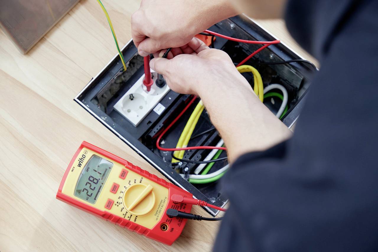 En person använder ett multimeter för att kontrollera elektriska anslutningar i ett vägguttag. Olika färgglada kablar är synliga.