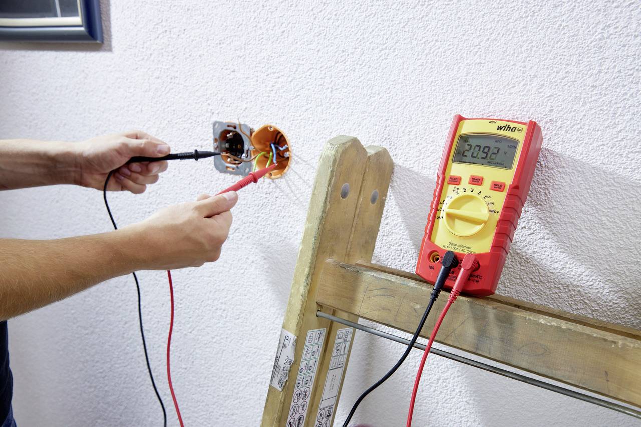 En person mäter spänningen i ett eluttag med ett digitalt multimeter som ligger på en trästege.