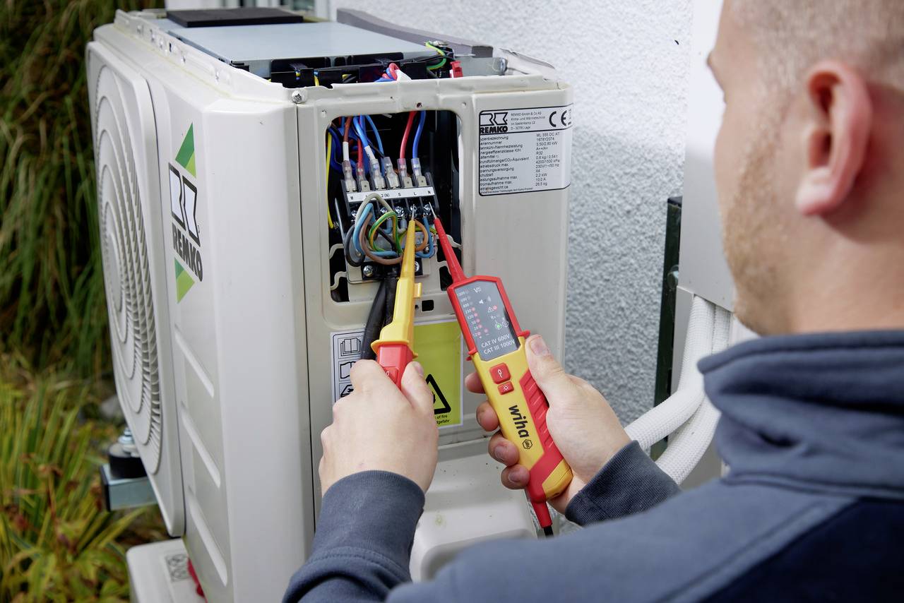En person kontrollerar de elektriska anslutningarna på en luftkonditionering utomhus med ett multimeter.