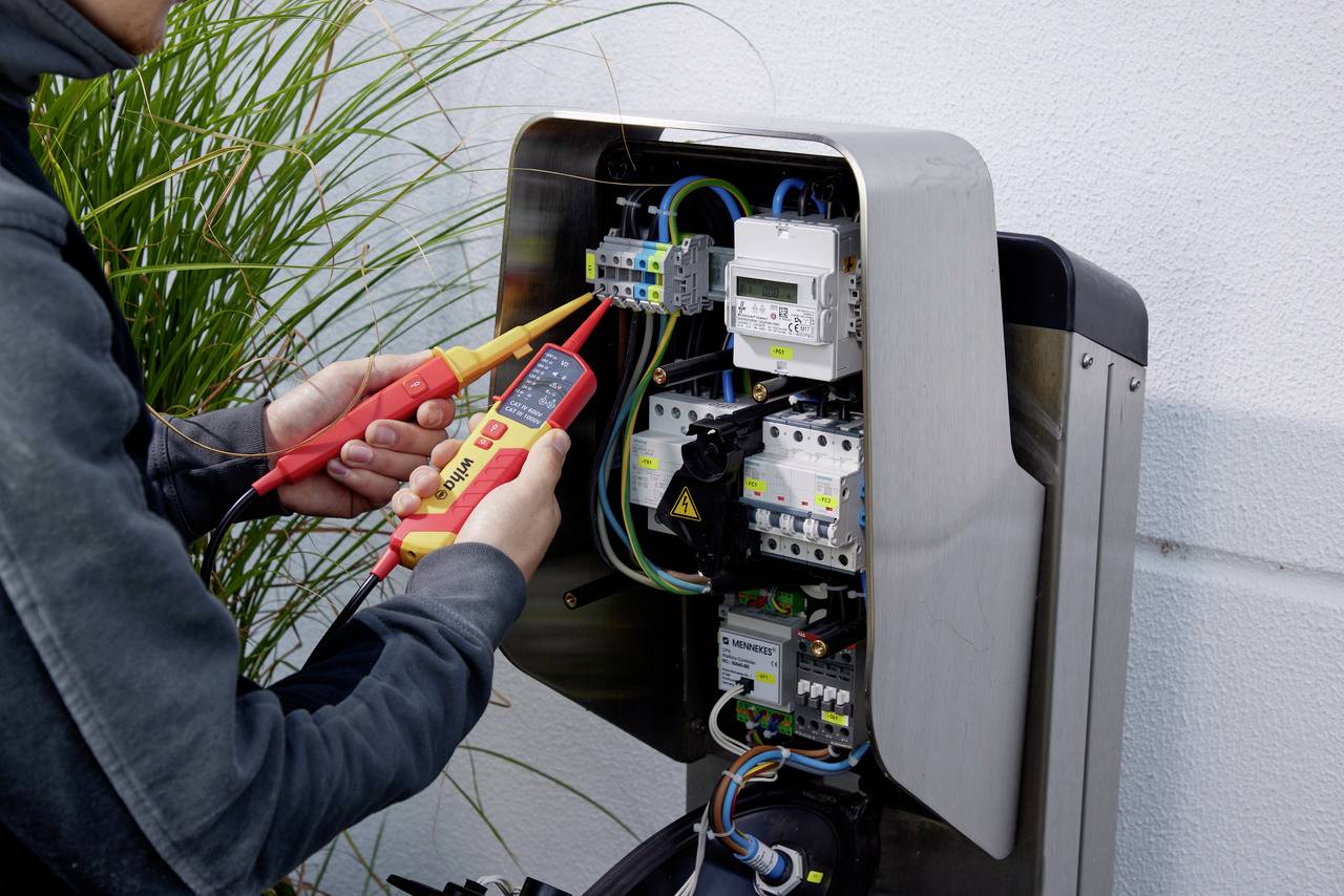 En person mäter elektriska anslutningar med ett multimeter i en öppen laddstation för att kontrollera säkerhet och funktionalitet.