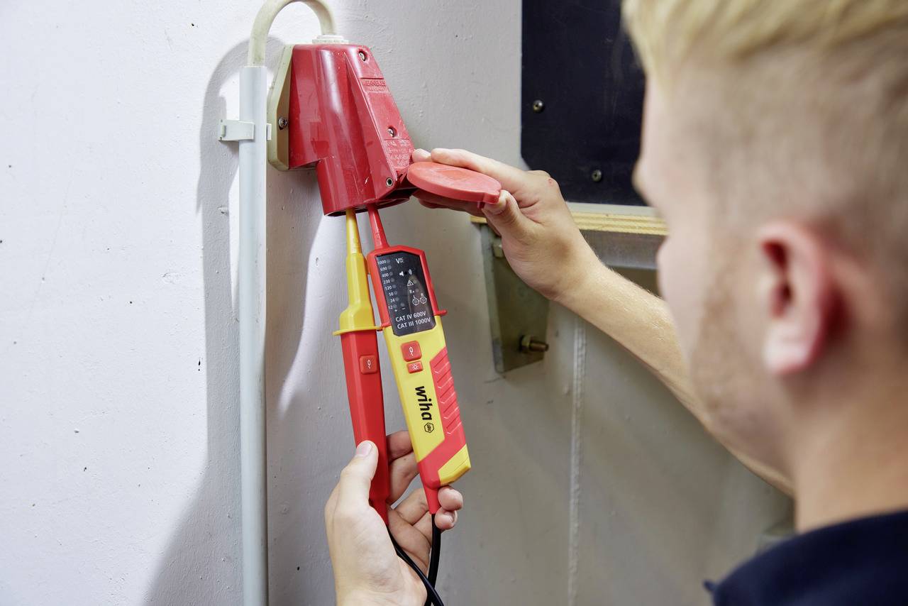 En elektriker kontrollerar den elektriska anslutningen i ett rött uttag med en spänningstestare.