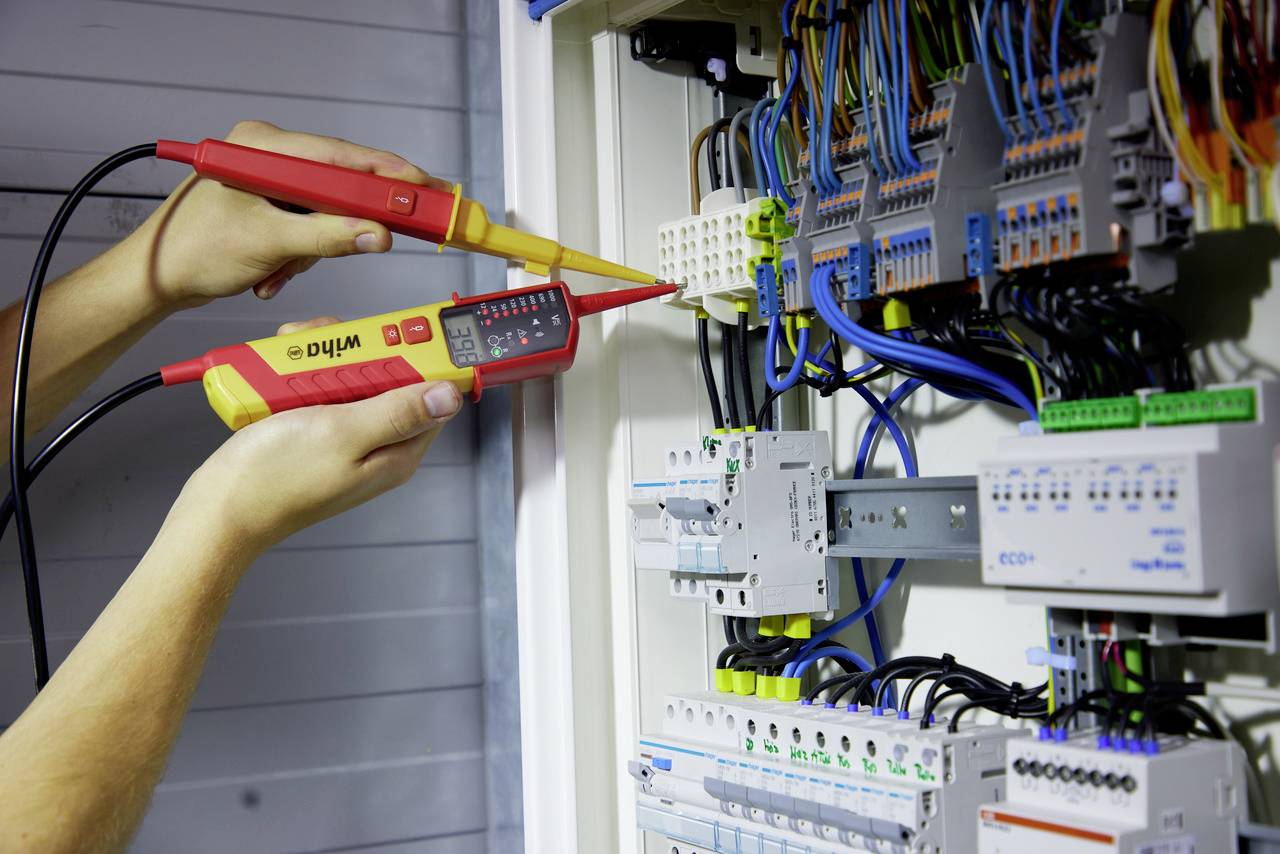 En person mäter elektrisk spänning i en kopplingsbox med en spänningstestare. Boxen innehåller olika kablar och säkringar.