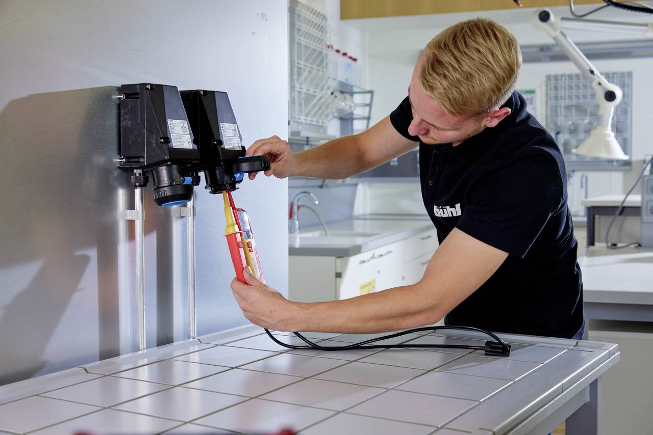 En man i arbetsklädsel testar elektriska anslutningar med en testapparat i ett laboratorium. I bakgrunden syns laboratorieutrustning.