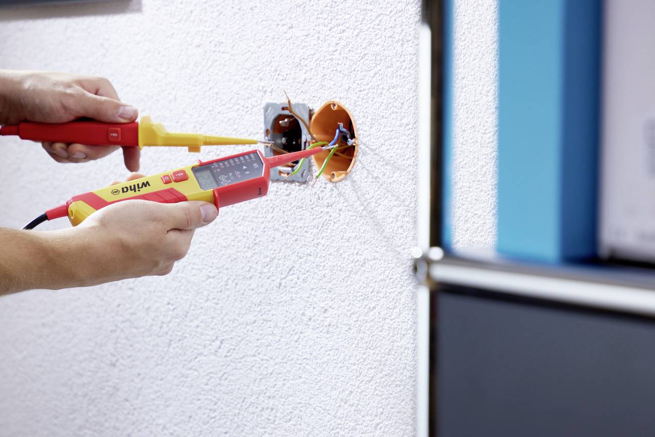 En elektriker arbetar med ett uttag och använder ett multimeter för att mäta spänning. Fokus är på handen och verktygen.
