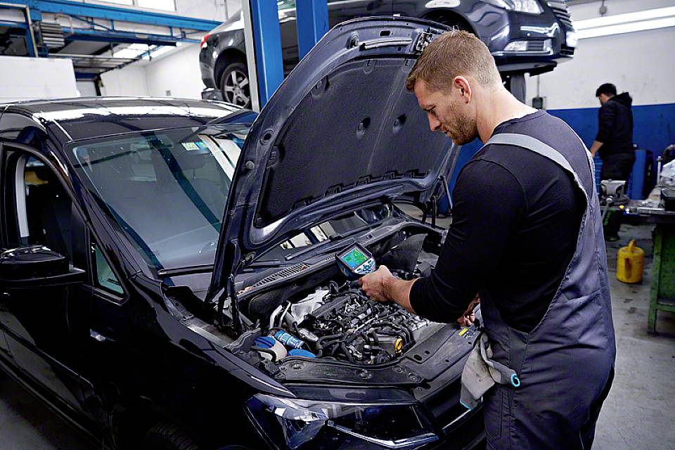 En mekaniker i en verkstad kontrollerar en bils motor med ett diagnosverktyg. Fler bilar syns i bakgrunden.