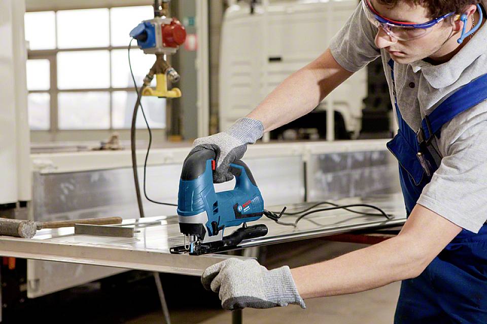 En arbetare i blå overall och grå handskar klipper ett metallstycke med en elektrisk sticksåg i en verkstad.