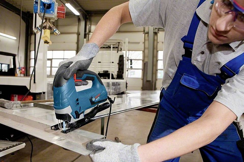 En arbetare i blå overall och grå handskar hanterar en elektrisk sticksåg för att såga ett stort metallstycke i en verkstad.