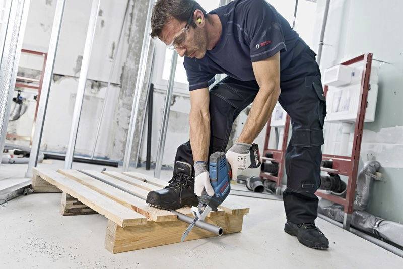 En man arbetar inomhus och använder en borrmaskin för att borra ett hål i ett rör på en träbalk.