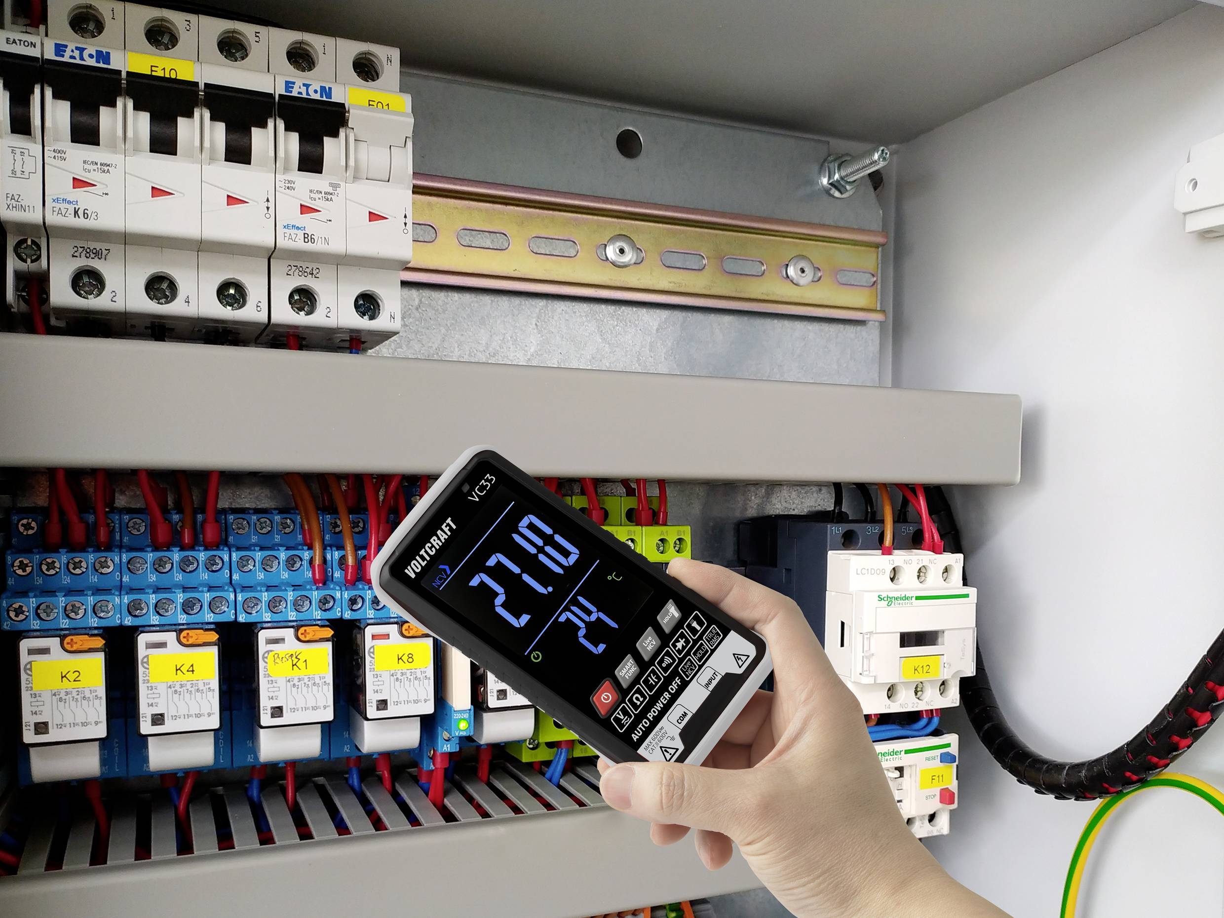 En hand håller ett digitalt mätinstrument som visar '27,10' framför en öppen elcentral med flera färgade kablar och säkringar.