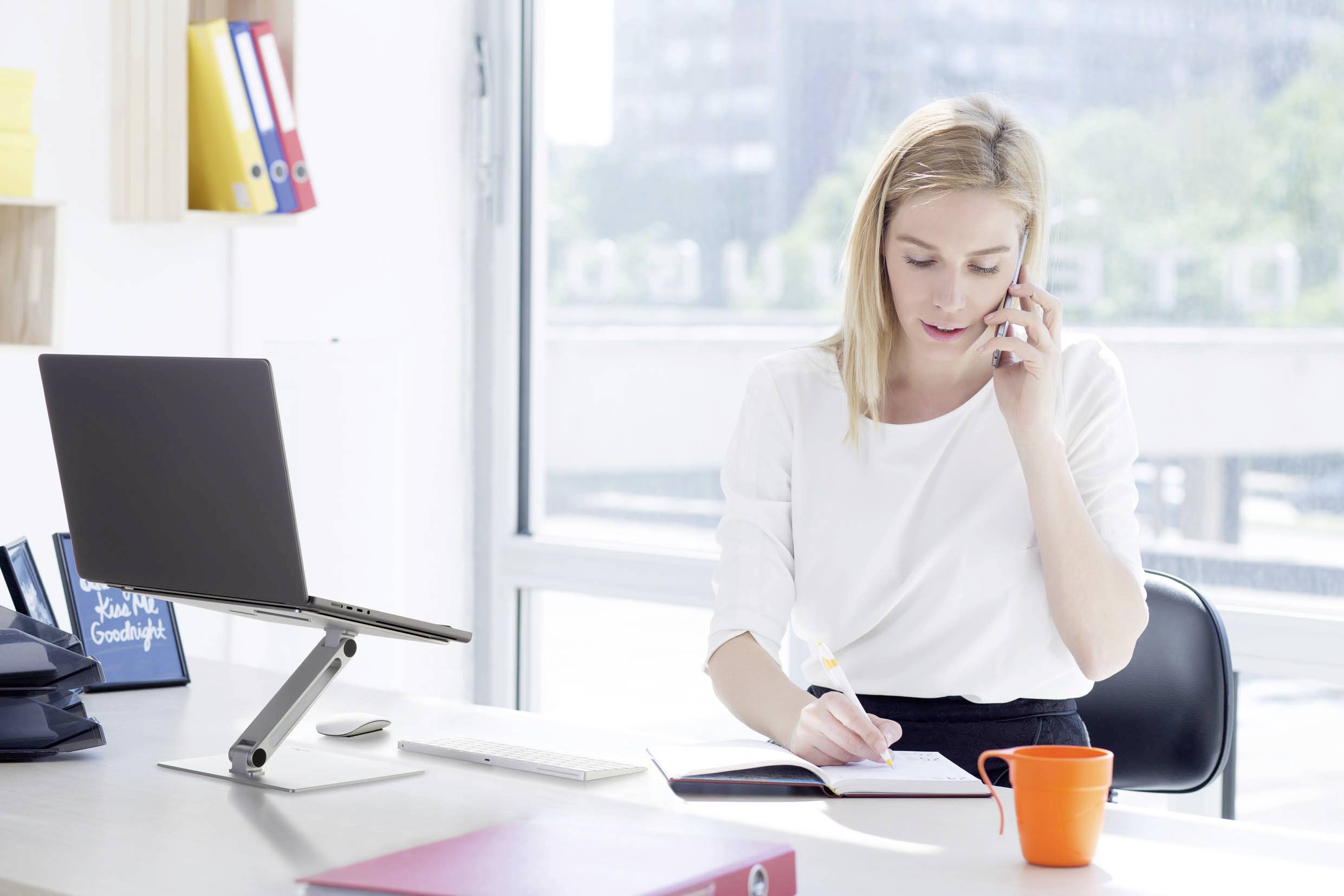 En kvinna sitter vid ett skrivbord, pratar i telefon och antecknar i en anteckningsbok. En bärbar dator står på ett laptopställ.